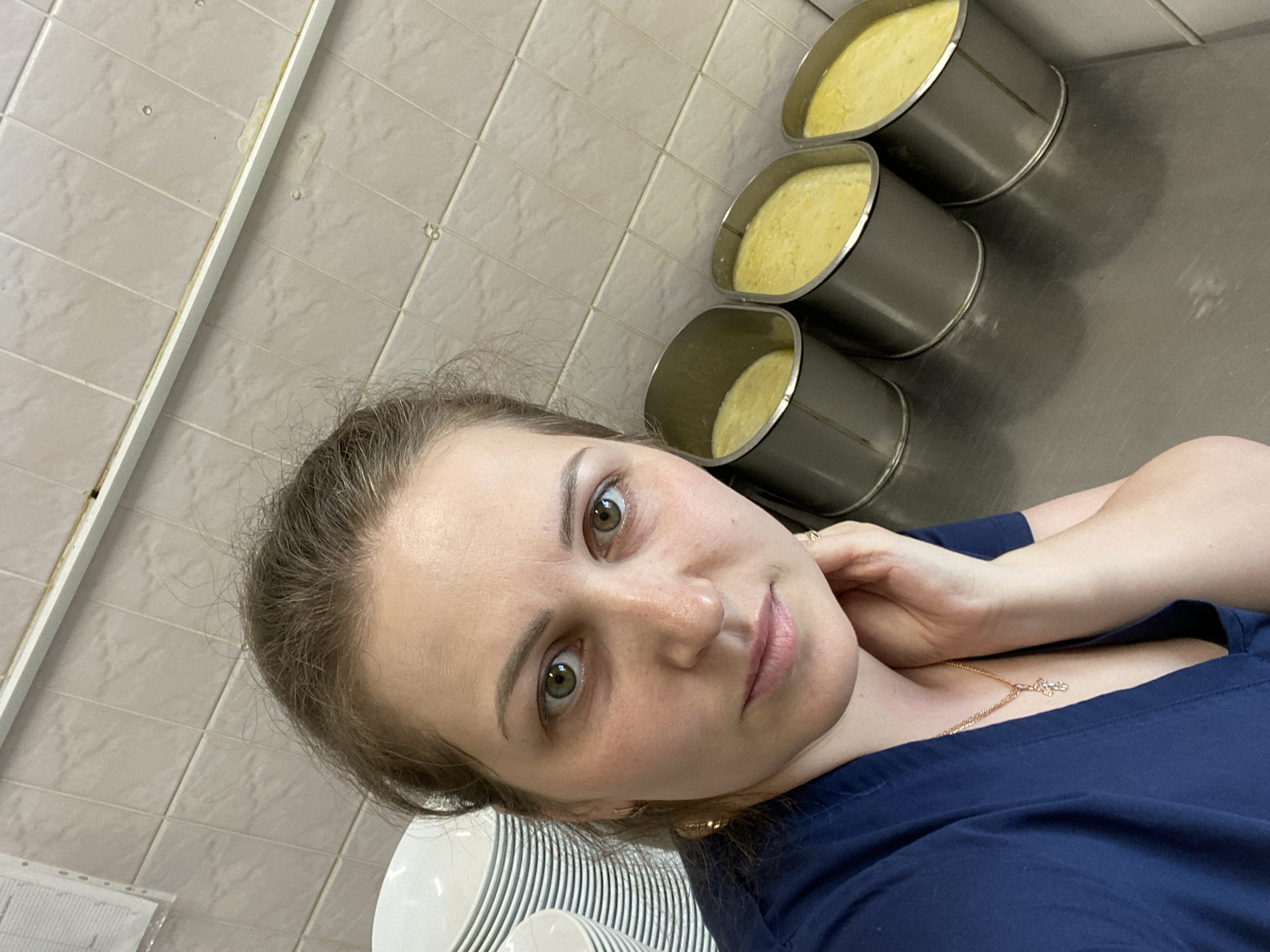 A woman is taking a selfie indoors in Марковский, Russia. In the background are three metal containers filled with a yellow substance, possibly food. To the left are a stack of white plates and a tiled wall. The woman is wearing a dark blue shirt and a gold necklace with a pendant. She is touching her neck.
