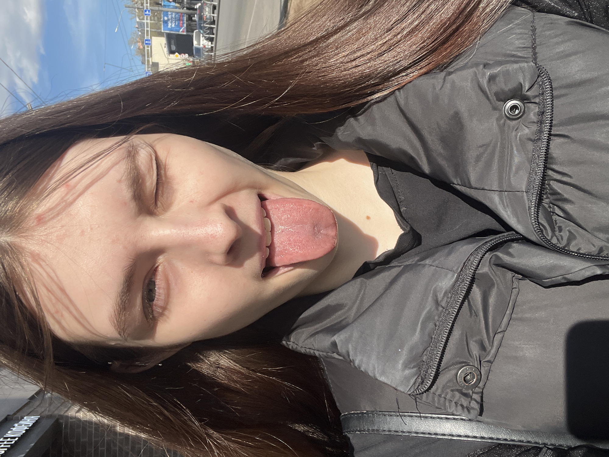 A young female with long, dark brown hair is captured in a close-up, rotated outdoor shot. She is sticking her tongue out and winking with one eye, conveying a playful and mischievous expression. Her skin is fair, with some minor blemishes visible on her forehead and cheek. She is wearing a black puffer jacket with a visible zipper and metal snap details, and a dark shirt underneath.
The background, out of focus, reveals an urban setting under a clear, bright blue sky with some clouds. Sunlight is strong, casting distinct shadows on her face and clothing, and highlighting her hair. Visible in the background are buildings, streetlights, and power lines. Several signs with text are discernible: a blue sign with "70%" and "POCCHЯ" (Russia) in white Cyrillic letters, and another sign below showing "COFFEE KODEHH" (likely part of a café name like "Coffee Copenhagen" or similar). The scene suggests a sunny day in a city, consistent with the given location context of Saint Petersburg, Russia.