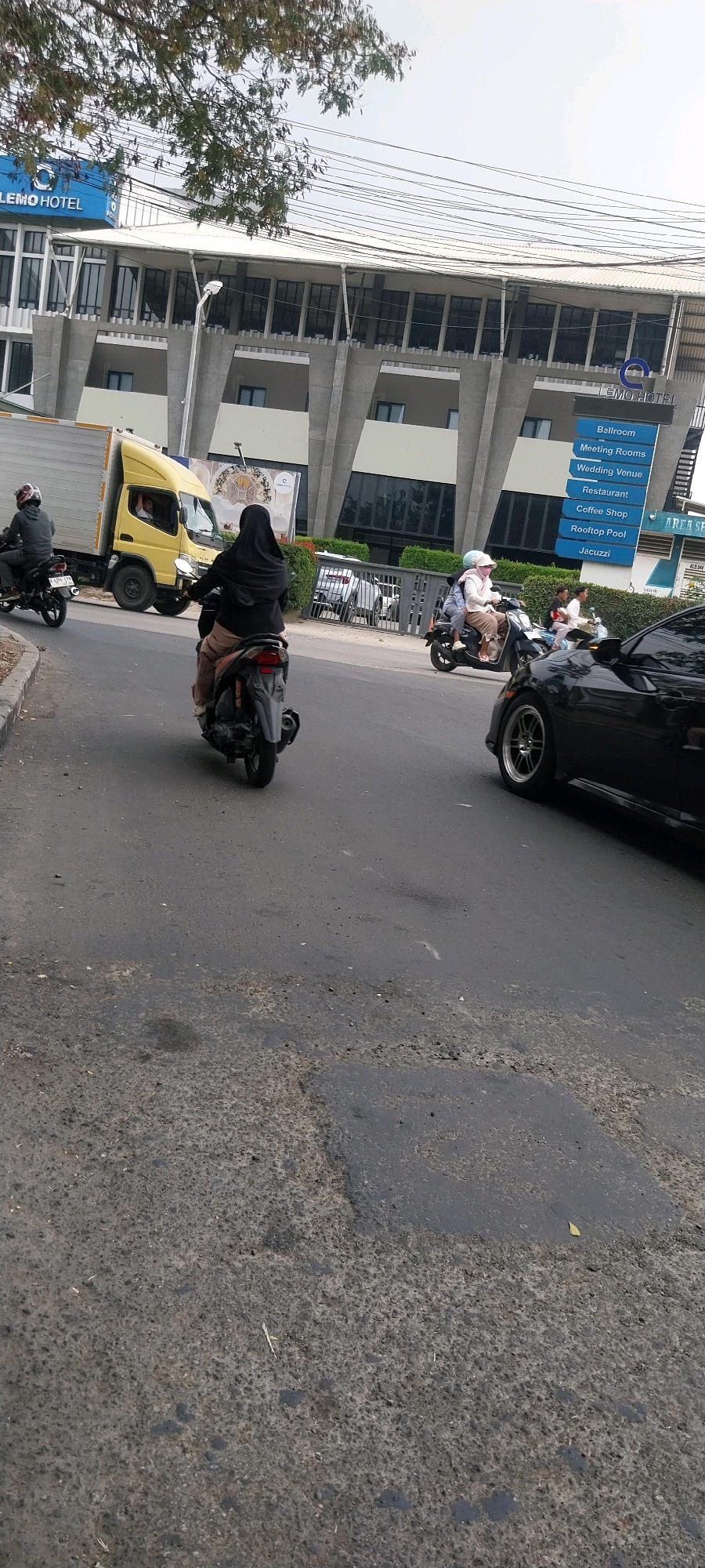 In a street scene in Kelapa Dua, Indonesia, a woman wearing a black hijab and tan pants rides a scooter. She is followed by a yellow truck. The truck's cab is visible with a man inside. Further down the road, two people on a scooter are seen, followed by a black car. In the background, a modern building with the sign "LEMO HOTEL" is visible, along with a blue sign listing hotel amenities. The street is paved with asphalt and shows signs of wear and tear. Trees with green leaves are present in the upper left corner of the image. The sky is overcast.