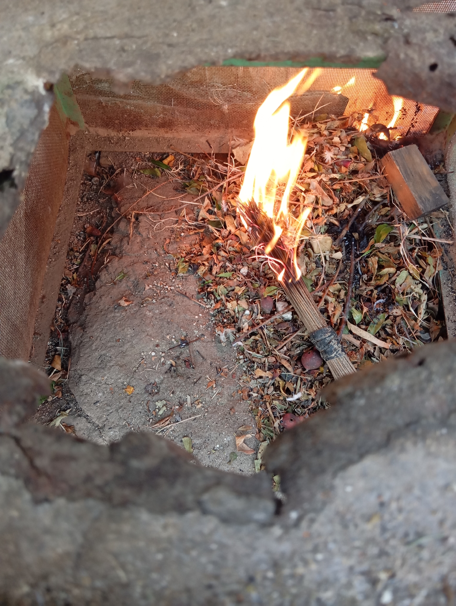 A torch made of wood and wrapped in black tape burns brightly in a shallow, makeshift enclosure. The enclosure appears to be constructed from wire mesh and is filled with dry leaves and debris. The ground around the enclosure is rough and appears to be concrete or dirt. The lighting suggests it is daytime, with shadows indicating a light source from above. The scene is located in the city of Hakale, Pakistan.