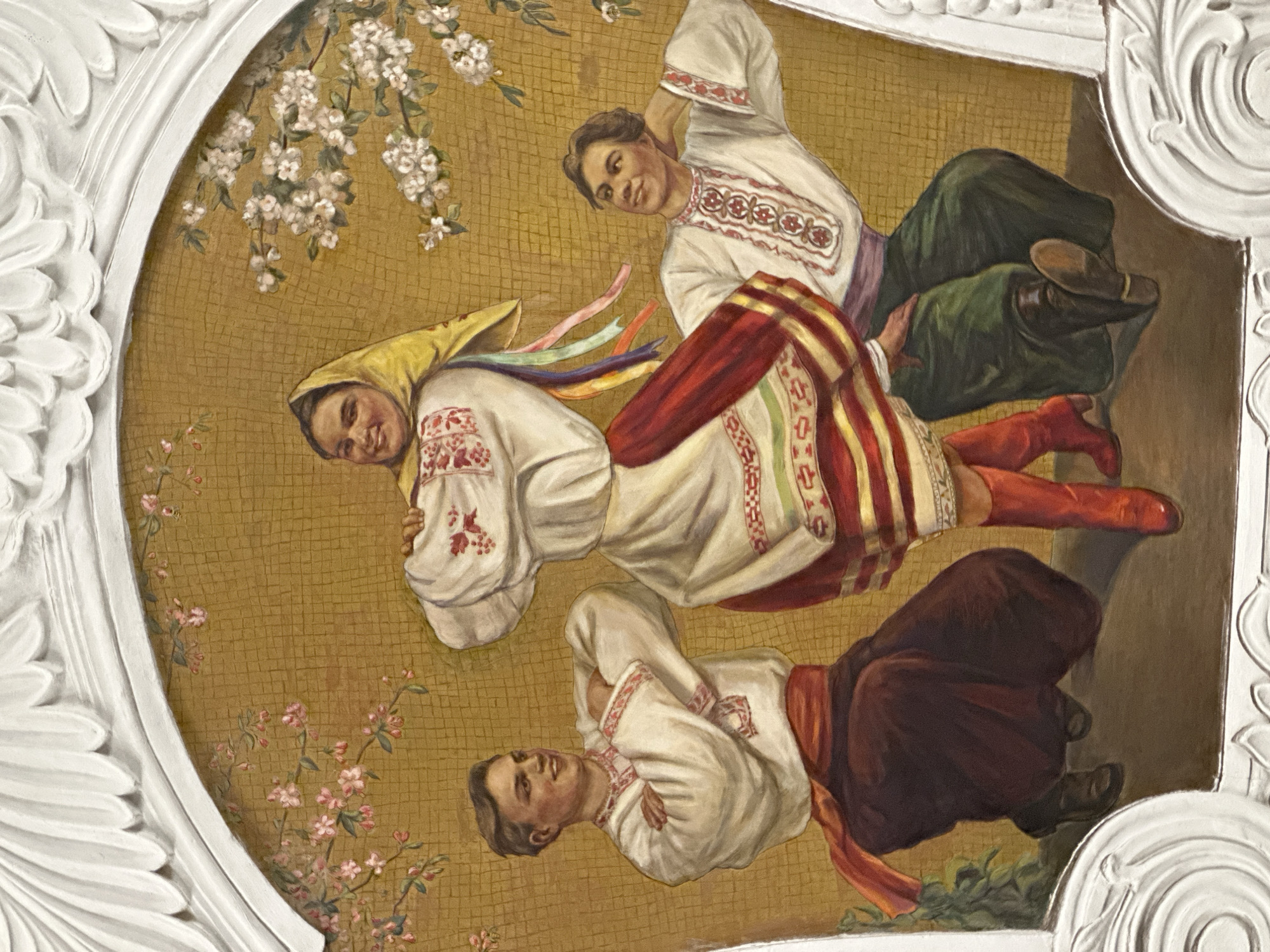 This is a painting of three people in traditional Ukrainian clothing, possibly engaged in a folk dance. The scene is set against a textured, golden background that resembles a mosaic. Floral elements, including branches with white and pink blossoms, are interspersed across the background, suggesting a spring setting.
The subjects are depicted in a dynamic, possibly celebratory, pose. Two individuals appear to be in mid-movement, with one person leaning back and arms outstretched, while another is positioned in a lower, bent posture. A third person is slightly above and behind the others, also appearing to be in motion. Their clothing features embroidered tunics, striped skirts, and vibrantly colored sashes and ribbons. One individual wears a yellow headscarf. The attire and the dynamic poses suggest a lively folk tradition.
The painting is framed by ornate white moldings, which are part of the architecture of the space where the artwork is installed. The overall impression is one of joy and cultural expression. There is no visible text in the image. The lighting suggests an indoor setting, likely with ambient light, as there are no strong shadows indicating a specific time of day. The emotions conveyed are positive and energetic, suggesting festivity or community gathering. The location context of Moscow, Russia, hints at the cultural context of the artwork, potentially showcasing Ukrainian traditions within a Russian setting.