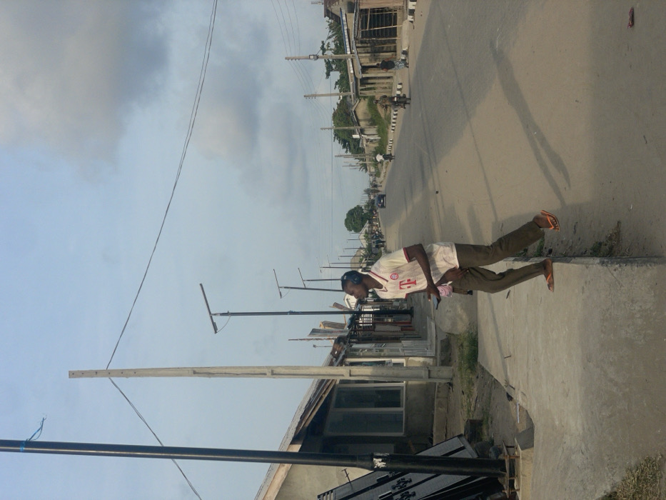 This image depicts a street scene in the city of Mosafejaw, Nigeria, under an overcast sky. In the foreground, a young man is positioned on the edge of a concrete sidewalk, facing left and looking upward. He wears a white polo shirt with a red collar and "T" logo, dark trousers, and orange flip-flops. Black over-ear headphones are on his head, and he holds a smartphone in his left hand. His body is angled, and his left foot is on the sidewalk while his right foot is suspended just above the dirt shoulder, casting a long shadow onto the street.
To the man's left, a black metal pole with a blue cord attached rises in the immediate foreground, alongside a lighter, bare utility pole. Behind these, a row of single-story buildings or stalls with varied facades, including corrugated metal and glass windows, line the left side of the street. Multiple black streetlights, featuring solar panels, are visible along this side, extending into the distance.
The paved road, appearing to be asphalt or tar, stretches into the background, flanked by dirt shoulders. On the right side of the road, more buildings of varying construction materials are present, along with several utility poles supporting power lines. Green foliage is visible further down the street.
In the mid-ground, a light-colored car is seen driving away from the viewer, and two individuals on motorcycles are also observed heading in the same direction. A solitary figure walks on the right shoulder further down the road. The overall environment suggests a semi-urban or suburban area with ongoing development and daily activity.