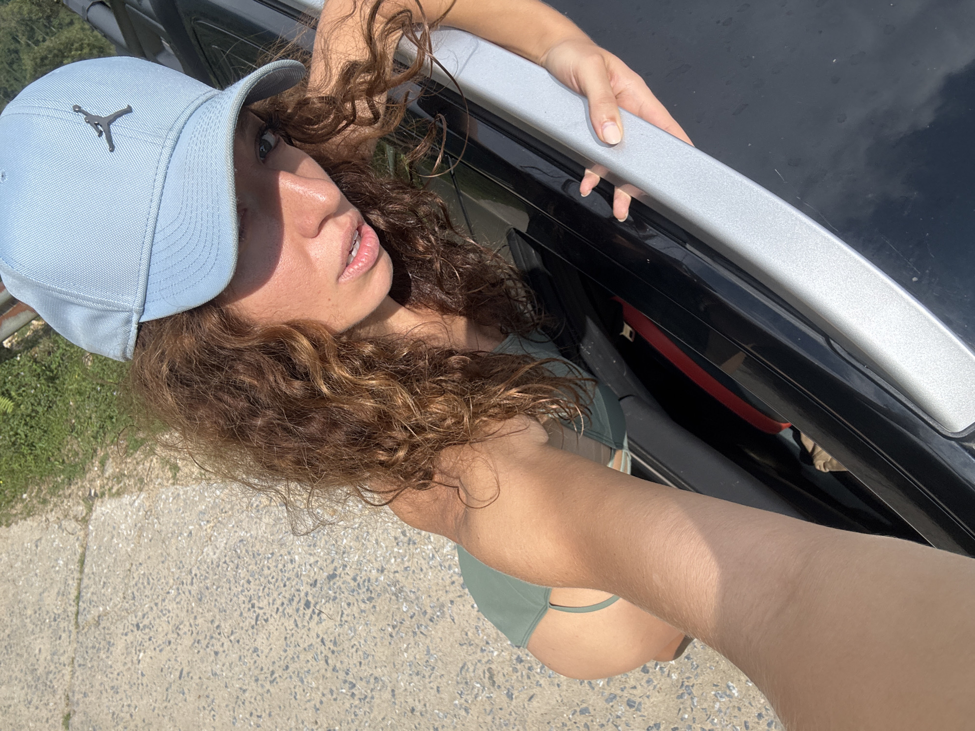 The image depicts a woman with curly brown hair wearing a light blue Jordan brand baseball cap and a green bikini. She is leaning out of the window of a black car, holding onto the silver roof rack. The background includes a road or pavement and some greenery. The location is given as Suối Giàng, Vietnam.