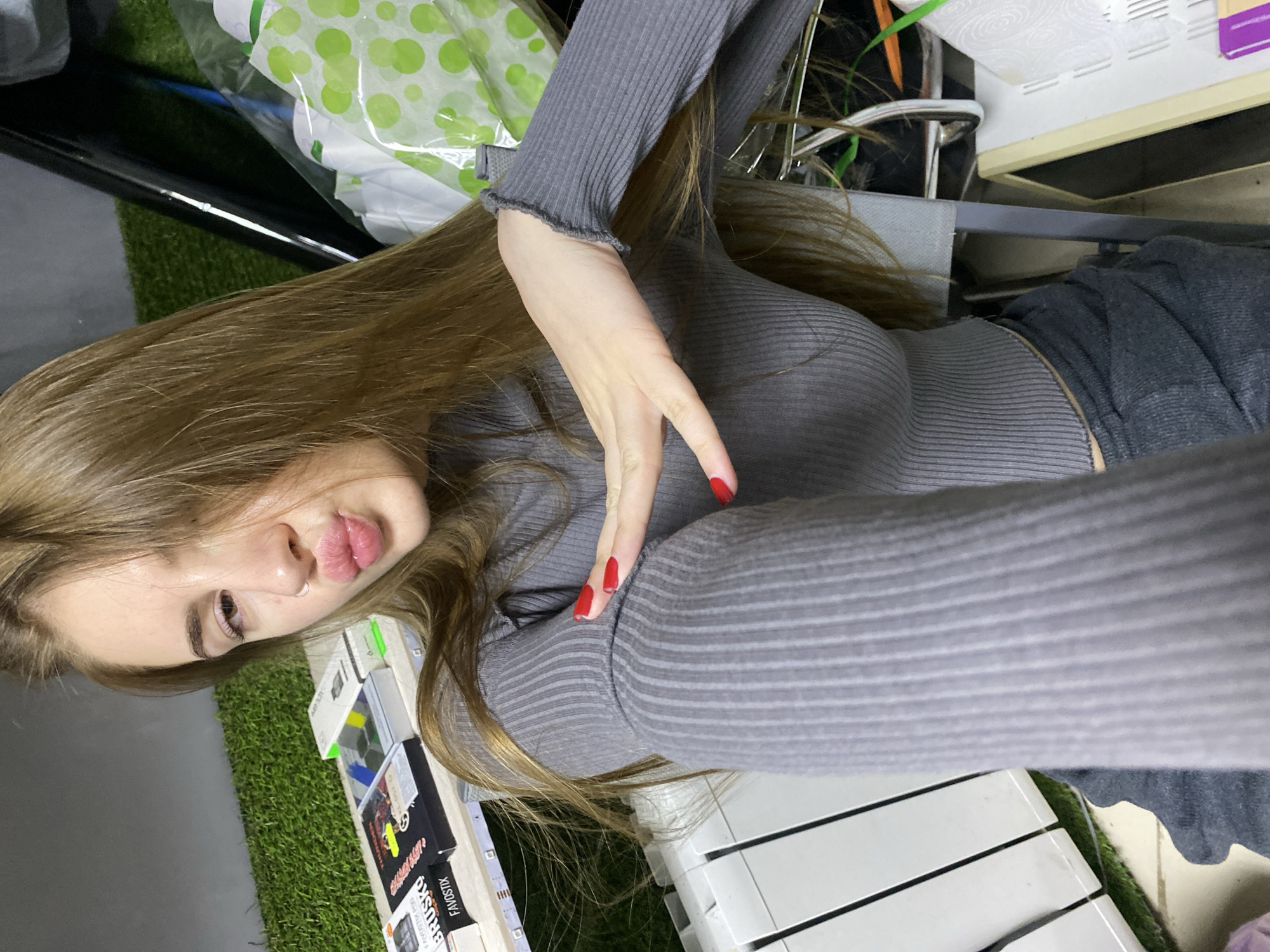 A young woman with long, blonde hair and a nose ring is taking a selfie. She is wearing a grey, ribbed turtleneck top and dark grey pants. Her lips are pursed, and she is holding her left arm up, pointing to her bicep with her right hand. Her fingernails are painted red. She is standing in what appears to be a room with artificial grass on the floor and various items on shelves behind her, including what look like electronic cigarette boxes. The background is cluttered and includes a white radiator. The lighting is dim. No specific landmarks in Dyakovo, Russia are identifiable in this image.
