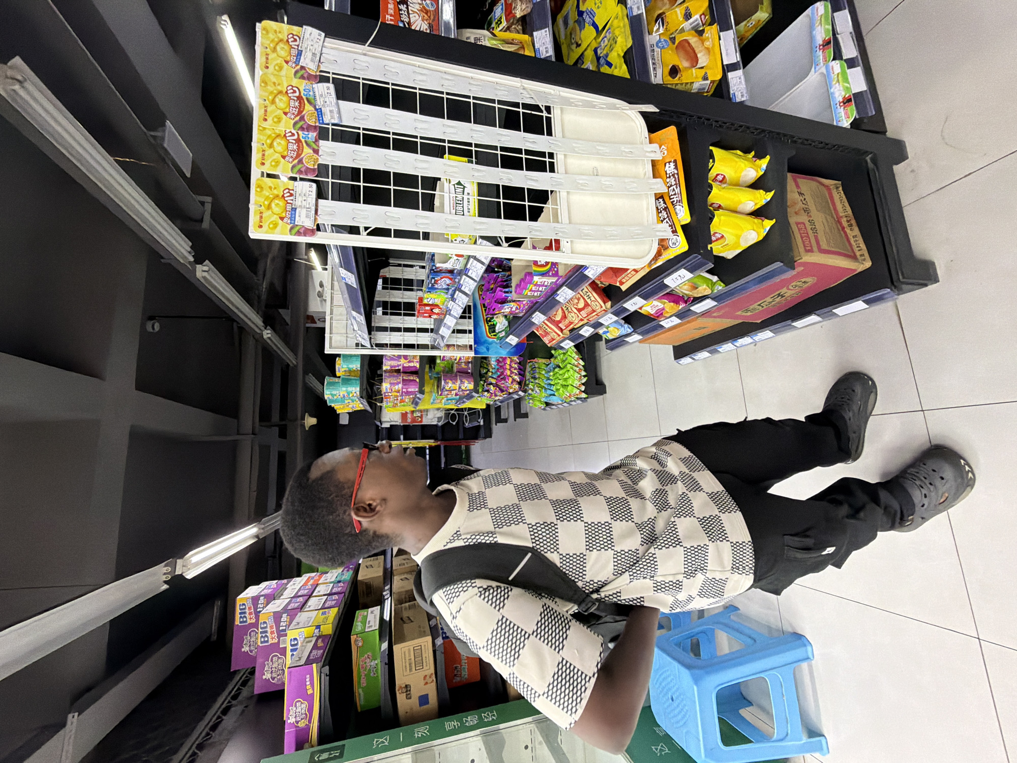 In the city of Jiangjin, China, a young Black male with short, dark hair and glasses with red frames stands in a supermarket aisle, looking at products on a shelf. He is wearing a checkered black and white t-shirt, black cargo pants, and dark grey clogs. A black backpack is slung over his shoulders. In front of him is a bright blue plastic stool. The shelves are stocked with various food items, including packaged snacks and drinks. The store appears to be well-lit with fluorescent lighting.