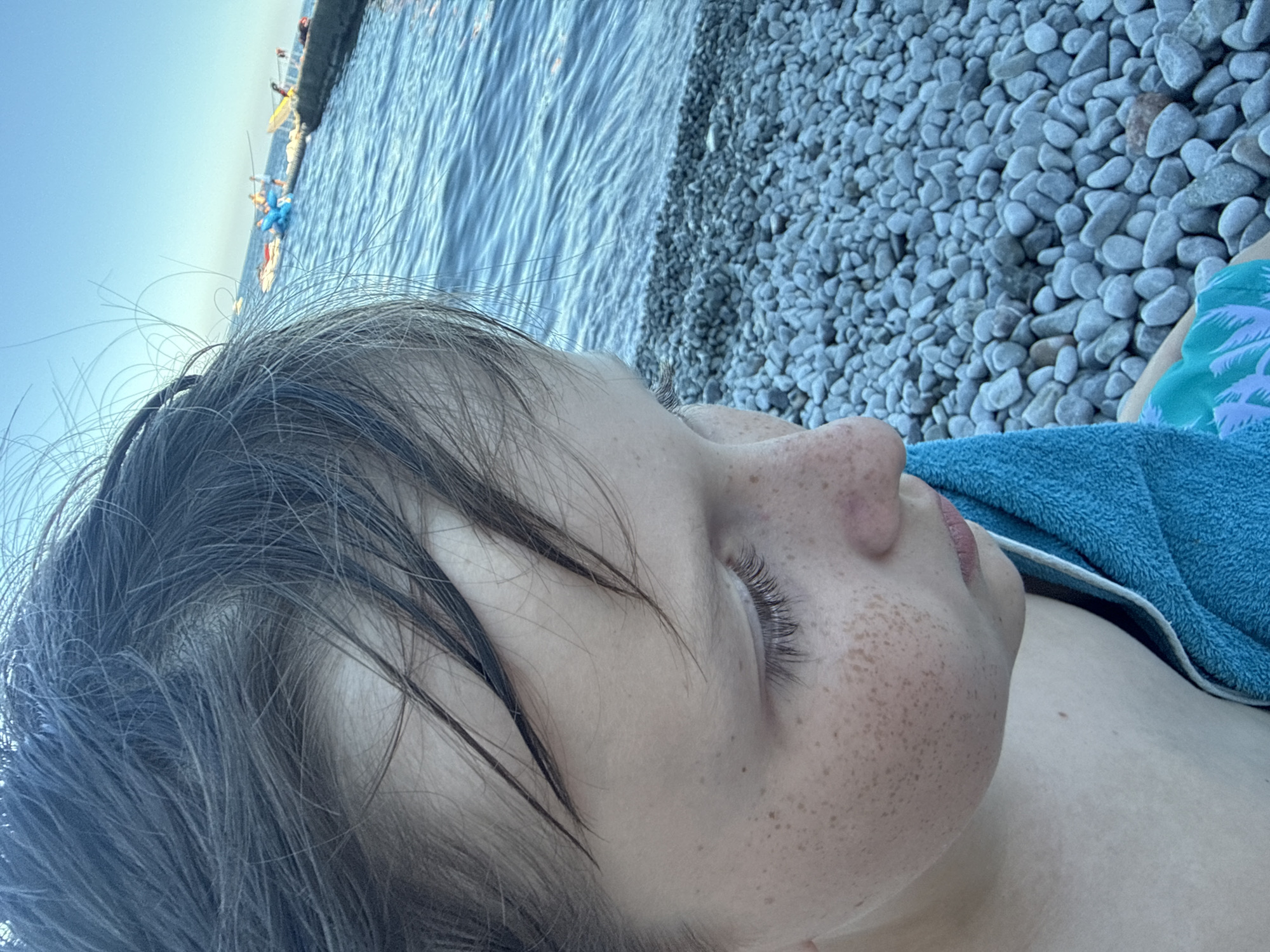 A close-up, over-the-shoulder shot of a young person's face and head is captured against a backdrop of a body of water and a pebble beach. The subject's face is turned to the right, with their eyes looking downwards. They have fair skin with a scattering of freckles across their cheeks and nose, and their dark hair is wet and disheveled, falling across their forehead. A bright teal towel is draped around their neck and shoulder.
In the background, the clear blue water of the sea stretches out to the horizon. Along the water's edge, a breakwater or pier is visible, with several small boats and what appear to be people on it. The beach in the foreground is covered in smooth, grey pebbles of varying sizes. The sky is a bright, clear blue, suggesting a sunny day.
This scene is consistent with the coastal environment of Partenit, Ukraine, a resort town located on the Crimean Peninsula known for its pebble beaches and the Black Sea.