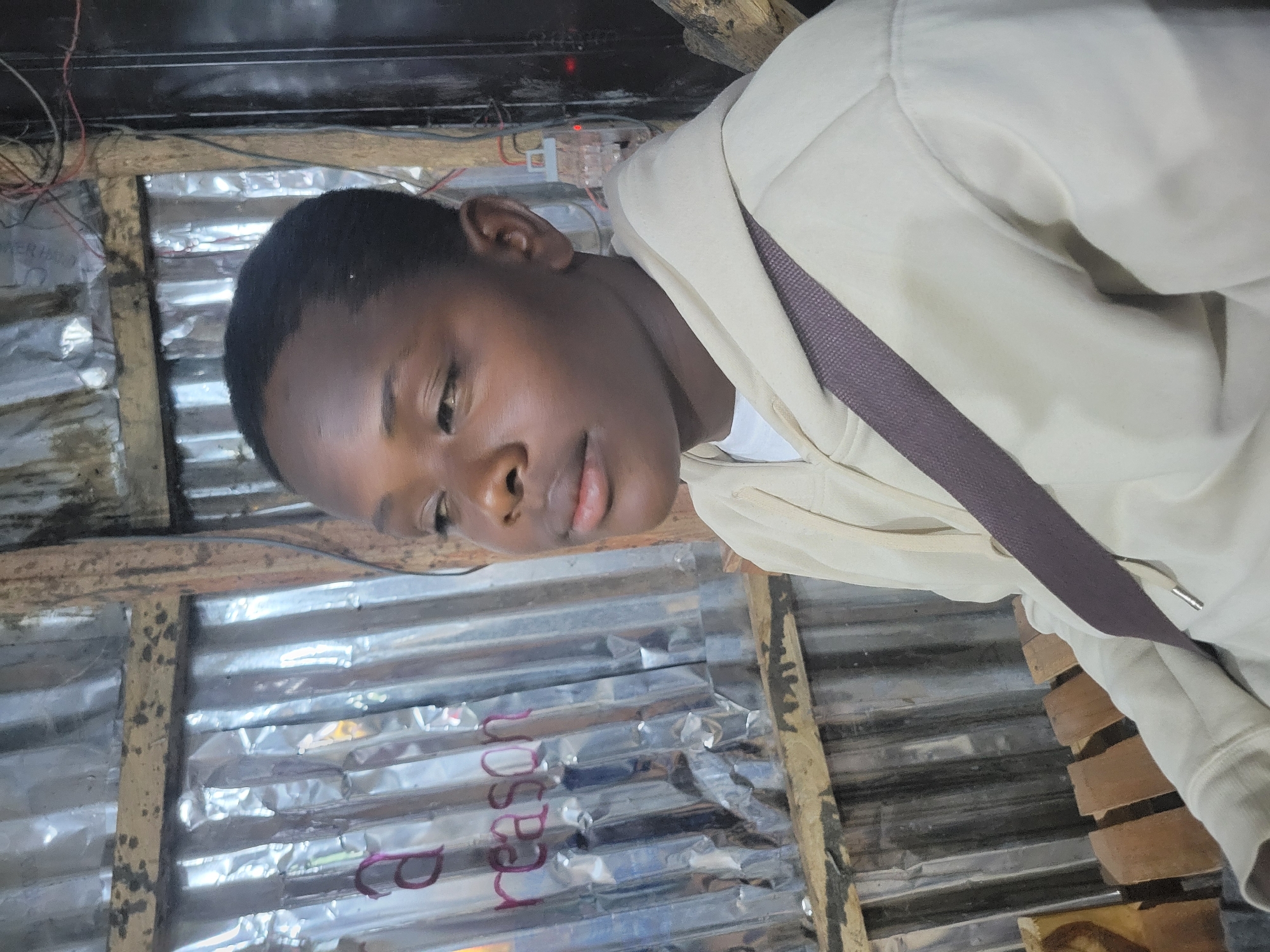 A young Black individual is pictured in a medium shot, facing left and looking away from the camera. They have short hair and are wearing a light-colored hooded sweatshirt with a dark strap crossing their chest. The background consists of corrugated metal sheeting, wooden planks, and some visible wiring. The lighting appears to be artificial and somewhat dim. There are some purple letters visible on the metal sheeting behind the individual, appearing to spell out "a reason". The setting appears to be outdoors, possibly in a market or makeshift structure. The location is identified as Durumi, Nigeria.