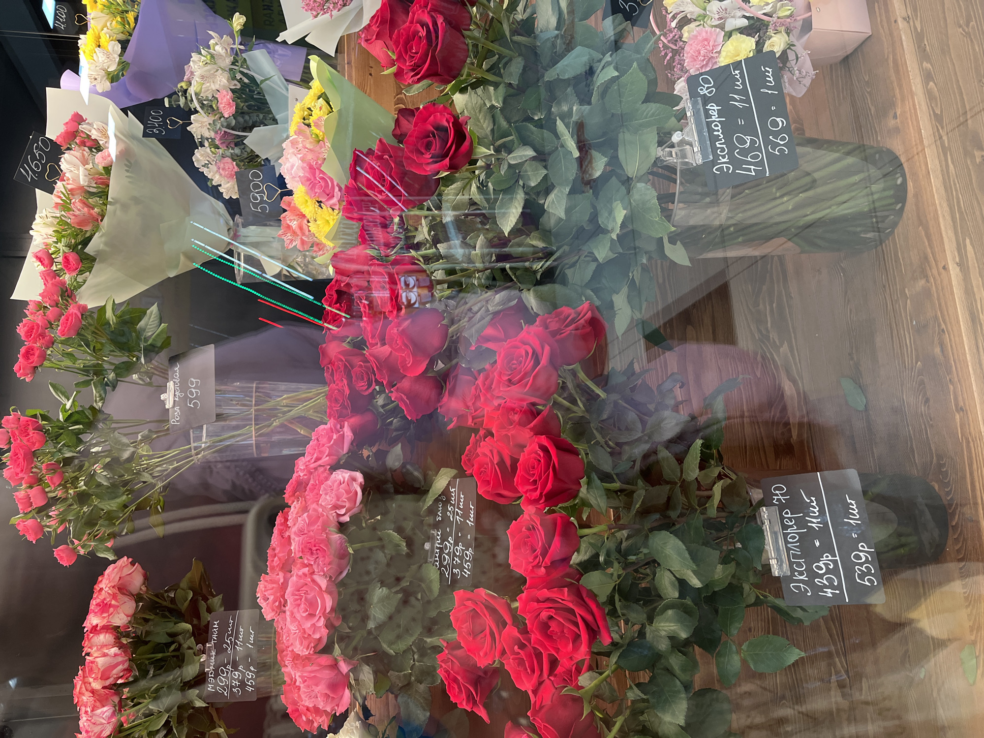 The image shows a flower shop window display. There are bouquets of flowers arranged inside, predominantly roses in various shades of red and pink. Some bouquets are wrapped, while others are in vases. Attached to some of the bouquets are price tags with Russian text, indicating prices for different quantities of flowers. The background is a wooden surface, likely part of the shop interior. The reflection on the window partially obscures the view, suggesting the presence of an external environment, although details are unclear. The text on the price tags suggests a Russian-speaking location, likely Petropavlovsk-Kamchatsky, Russia, given the instruction.
