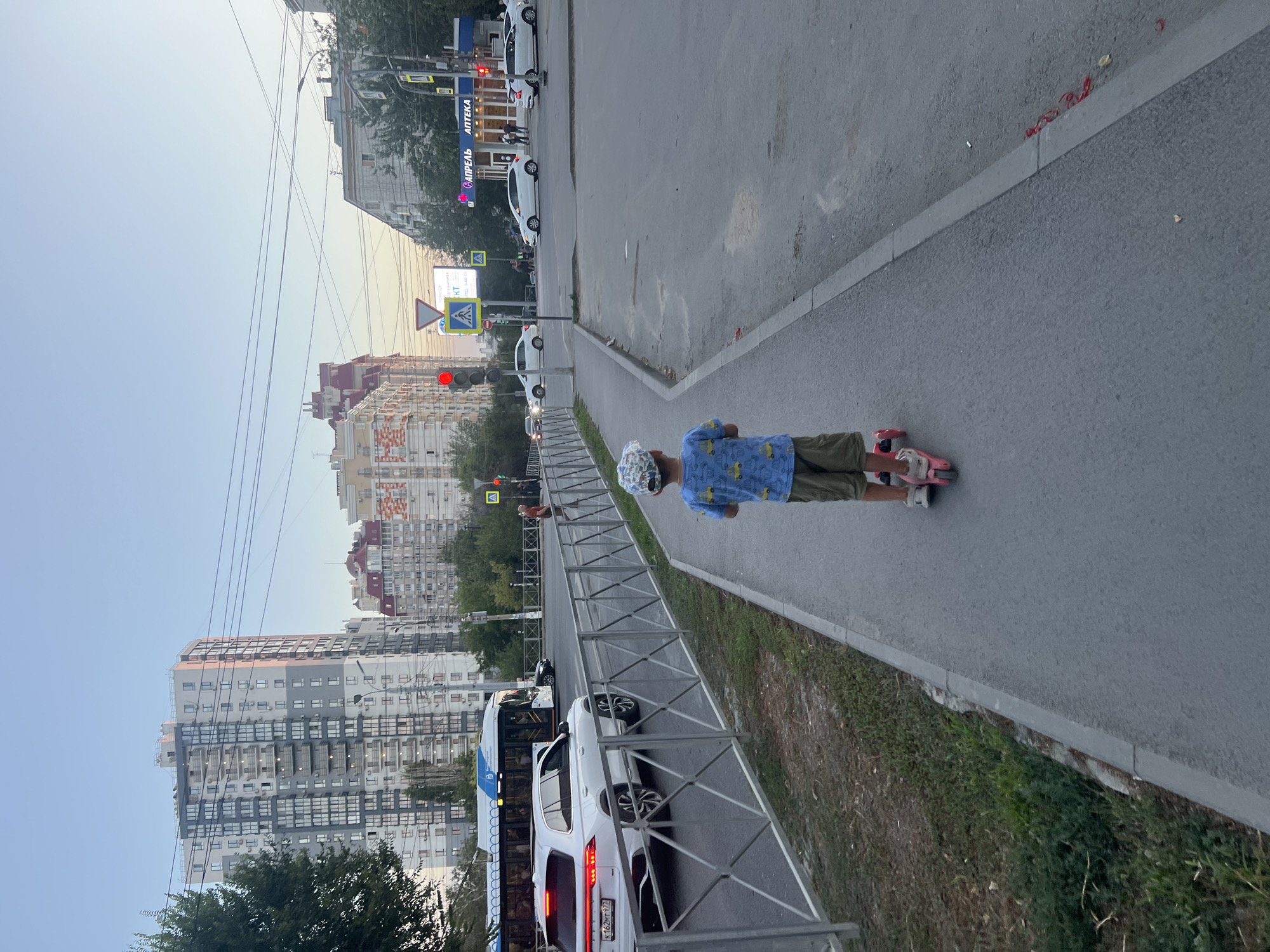 In Volgograd, Russia, a young boy wearing a blue t-shirt with a yellow car print and a patterned cap is riding a pink scooter along a sidewalk. He is facing away from the camera, towards a street scene. The street features multiple lanes of traffic, a bus, and several cars. Tall apartment buildings dominate the background, with a clear sky overhead. Traffic lights and street signs are visible, indicating an urban environment. The boy is on the right side of the sidewalk, with a metal fence separating the sidewalk from the road. The overall scene depicts a typical urban setting during daylight.