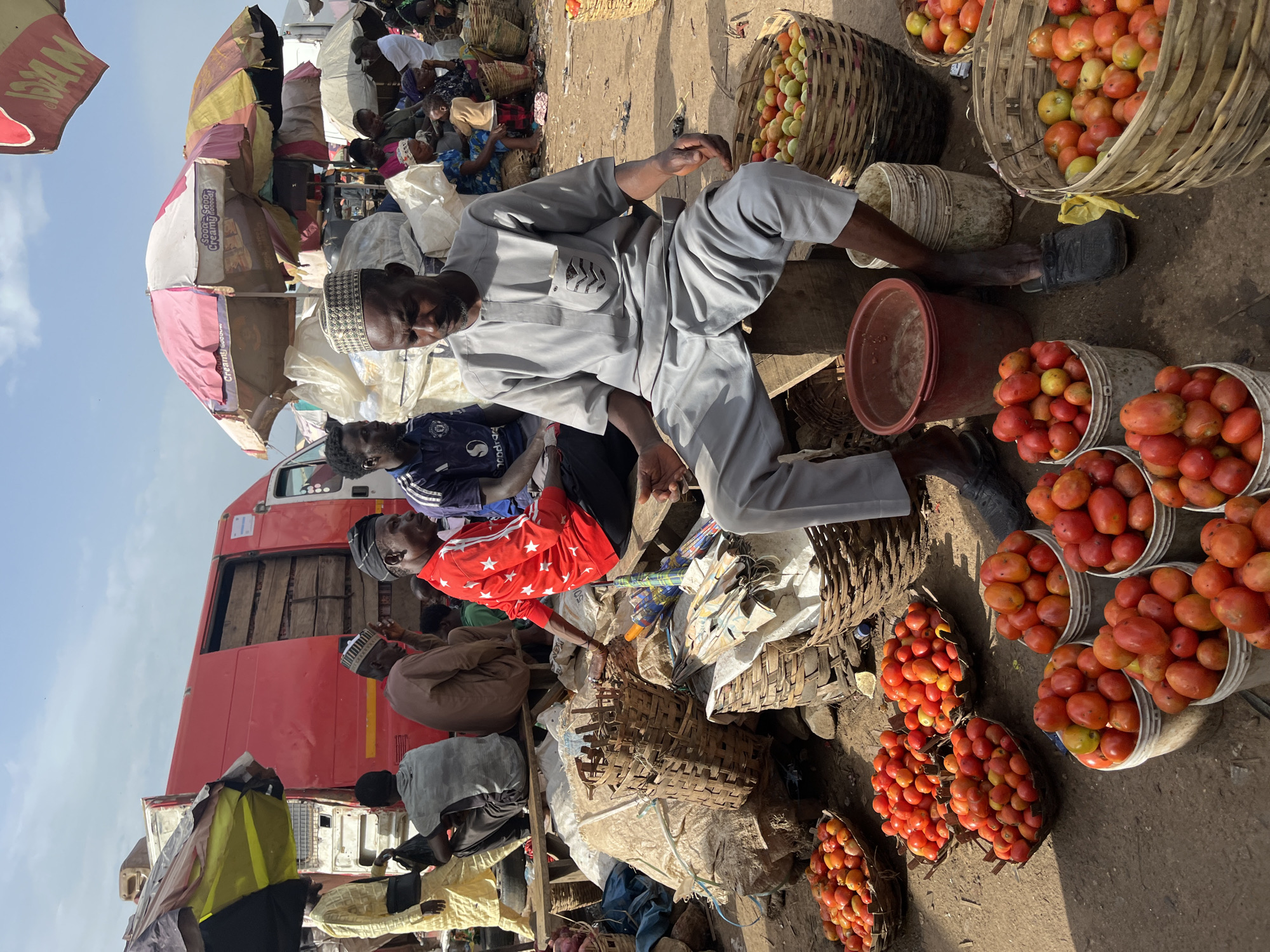 The image depicts an outdoor market scene in Rafin Par, Nigeria. In the foreground, a man wearing a gray cap and traditional attire is seated on a wooden stool, with baskets of tomatoes in front of him. He appears to be engaged in conversation or observing the surroundings. Several other individuals are visible in the background, some seated and others standing, near a red truck and market stalls with umbrellas. The ground is dusty, typical of an outdoor marketplace. The overall atmosphere suggests a bustling commercial environment.