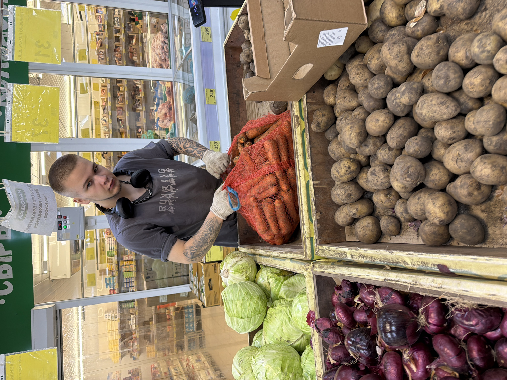 The image shows a man wearing a grey t-shirt, headphones around his neck, and gloves, standing in a grocery store in Chaikovsky, Russia. He appears to be reaching into a net bag of carrots. The carrots are located above heads of cabbage and red onions. A bin of potatoes is also beside the carrots. The aisles of the store are visible in the background, with refrigerated displays of meat and other goods. Price signs are seen throughout the photograph.