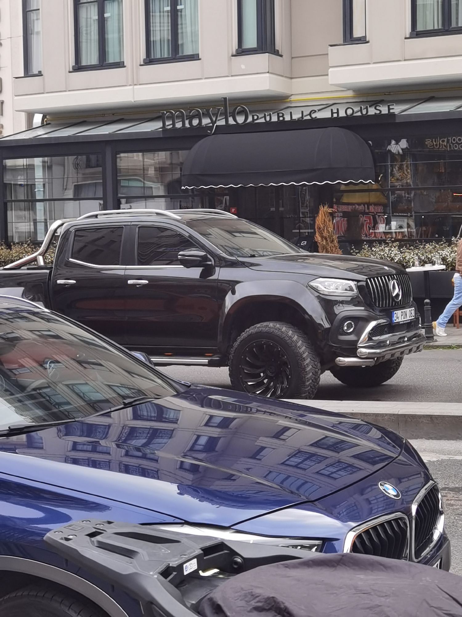 This daytime image captures a vibrant street scene in Beyoğlu, Turkey, featuring a commercial building, parked vehicles, and a glimpse of a pedestrian. The lighting suggests an overcast day, with soft reflections on the car surfaces.
In the foreground, the front of a dark blue BMW is visible, with its glossy hood reflecting the architectural details of the building across the street. A black, covered object, possibly a motorcycle or scooter, partially obscures the BMW's lower section.
The central subject is a striking black Mercedes-Benz X-Class pickup truck, modified with off-road enhancements. It features a bold, vertically-slatted front grille bearing the Mercedes-Benz star, large knobby tires mounted on custom black wheels, and a chrome bull bar and side steps. A chrome roll bar is also visible in its truck bed. The Turkish license plate "34 PON 082" is clearly displayed on the front bumper.
Behind the vehicles, a multi-story building with large windows forms the backdrop. Below the building is a storefront with a black, scalloped awning. The business name, "maylo PUBLIC HOUSE", is prominently displayed in silver lettering above the awning. Through the large glass windows of the storefront, some interior details, including plants and what appears to be a sign with "20₺", can be seen. On the far right, a person wearing light-colored jeans and a brown jacket is partially visible, walking past the storefront. The scene depicts a typical urban environment with upscale vehicles and commercial establishments.