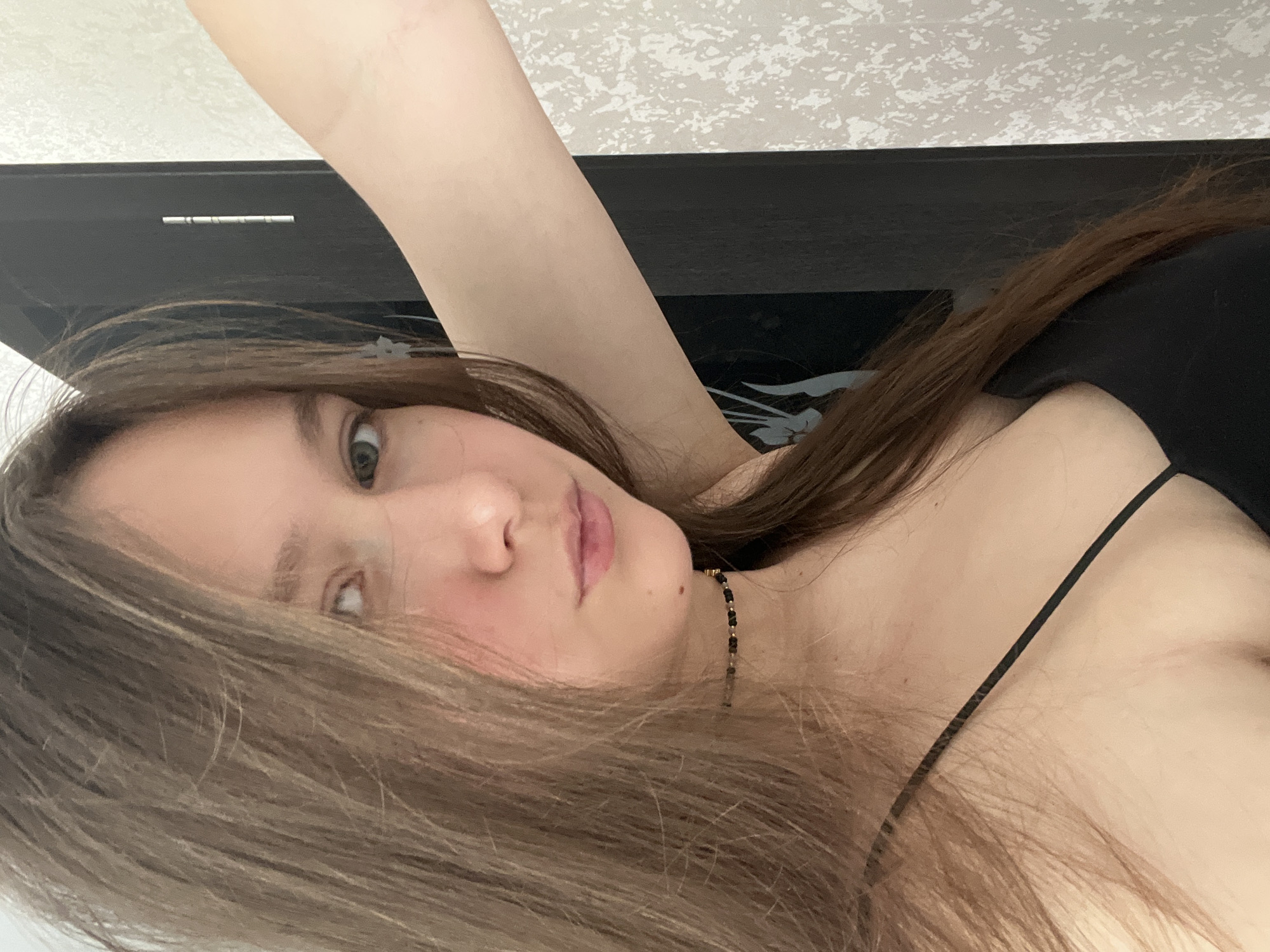 A young woman is seen in a close-up shot, possibly a selfie. Her hair is long and light brown. She wears a thin-strapped black top and a beaded necklace. The background includes a white patterned wall and a dark, possibly black headboard. No specific indicators of the city of Chaikovsky, Russia are visible.