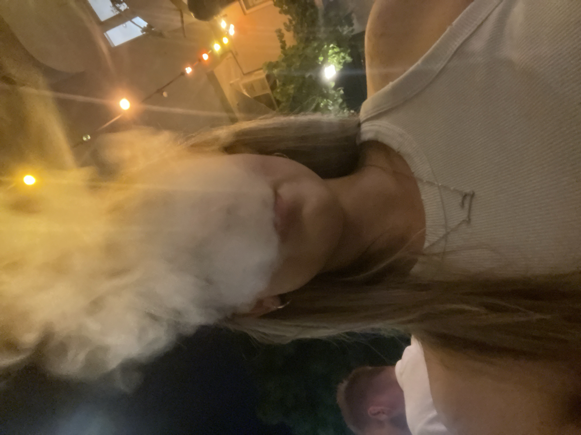 The image displays a female subject, primarily visible from the chest up, exhaling a large volume of white smoke that obscures the upper portion of her face. She has long, light-colored hair and wears a light-toned, ribbed tank top with a delicate silver-colored necklace featuring a small, indistinct pendant. A small hoop earring is visible in her left ear. To her left, partially visible, is the back of another individual's head and shoulder, wearing a white t-shirt. The background is dark, suggesting a nighttime setting, illuminated by numerous warm-toned string lights (yellow, orange, red) strung across the upper right. Indistinct architectural elements and some green foliage are also visible in the background. The scene takes place in the city of Unknown, Unknown.