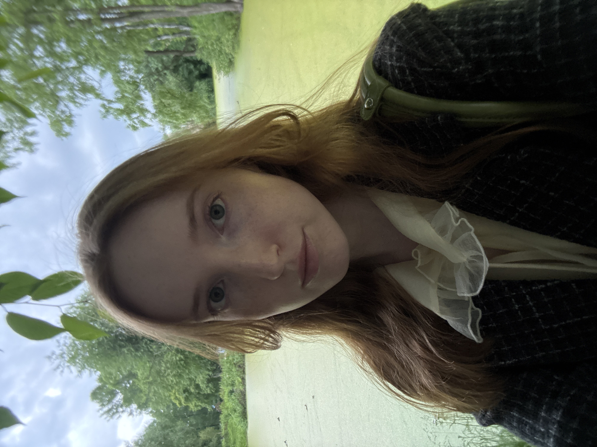 A young woman with red hair and blue eyes is taking a selfie outdoors. She is wearing a dark tweed jacket over a cream-colored blouse with a ruffled collar. A green strap, likely from a bag, is visible on her shoulder. The background features a body of water covered in green algae, surrounded by trees and foliage. The sky is overcast. The scene appears to be in a natural setting, possibly a park or a pond area within a city. The image is taken at a close-up to medium shot, focusing on the woman's face and upper body.