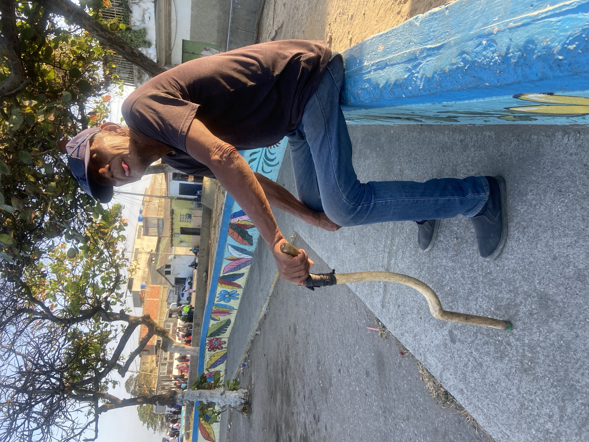 A man in Higuerote, Venezuela, leans over a blue concrete barrier. He wears a dark t-shirt, blue jeans, and black shoes, with a baseball cap and sunglasses perched on his head. He uses a crooked walking stick to support himself. The barrier is adorned with colorful floral paintings. Buildings are visible in the background. A tree with dense foliage overhangs the scene.