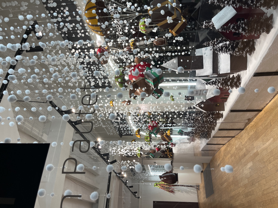 A retail display features hanging decorations: spheres resembling snow and various chocolate-themed objects like hot air balloons, gift boxes, and houses. The display is visible through a glass window, reflecting the interior of the store, including a wooden floor. The name "pâtisserie" is partially visible.