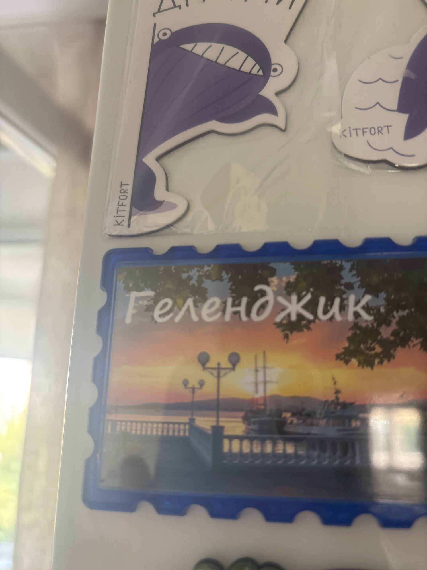 The image shows a close-up of several magnets on a white surface, possibly a refrigerator. One magnet features a stylized purple whale graphic with the text "KITFORT" visible. Another magnet displays a scene with a sunset over a body of water, a promenade with railings, streetlights, and a ship. White Cyrillic text, which appears to read "Геленджик" (Gelendzhik), is superimposed on this image. The background is bright and out of focus, suggesting natural light. The magnets are layered and appear to be slightly worn. There is no discernible indication of the city of Dubovoye, Russia in the image.