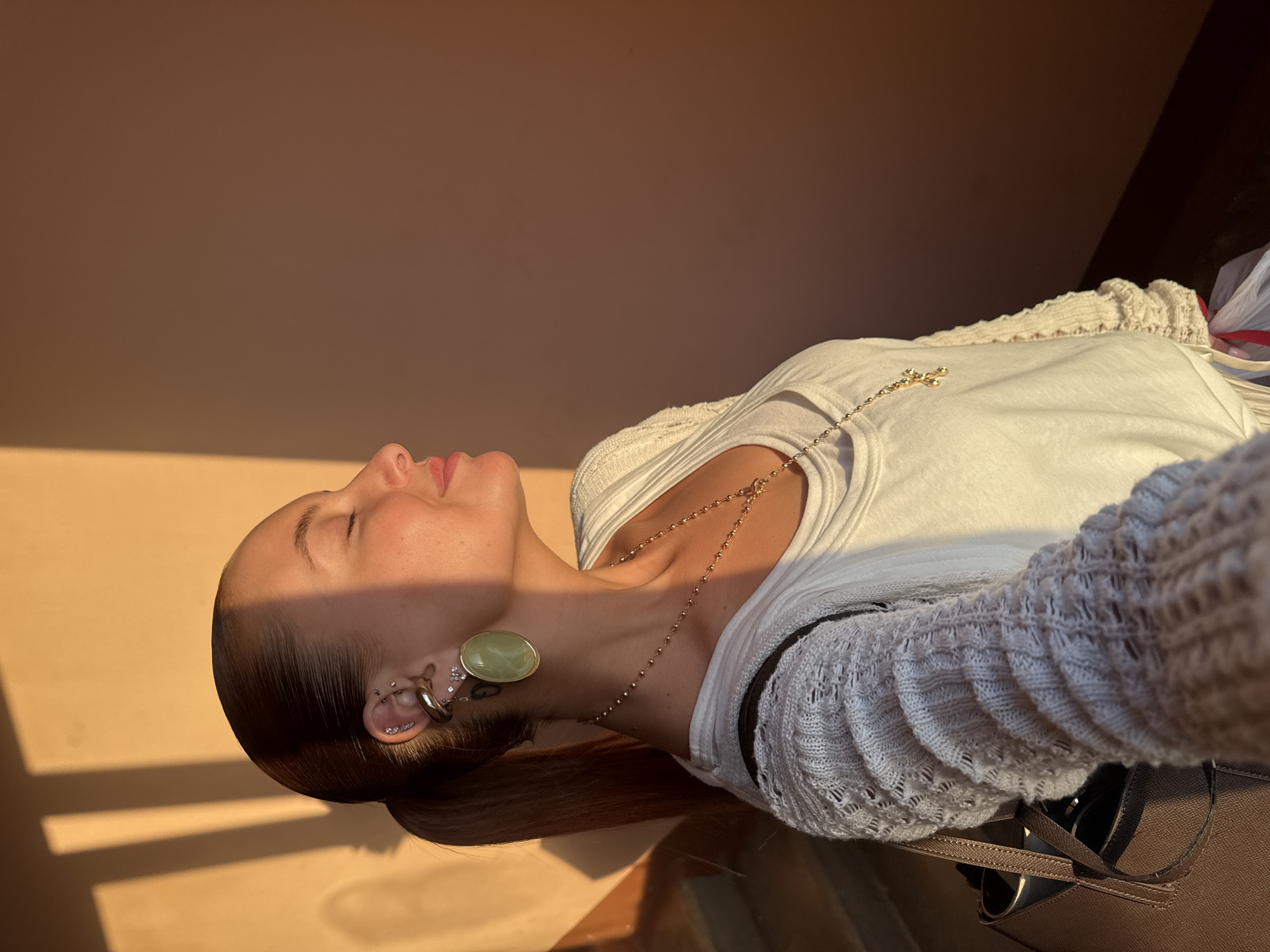 A woman with fair skin is lying on a flat surface in a room with golden light, possibly reflecting off a wall. She is facing upwards with her eyes closed. She is wearing a white t-shirt and a light-colored knitted sweater with a distinct pattern. A gold chain necklace with a cross pendant is visible on her chest. She is also wearing a large, light green earring in her left ear, along with other smaller earrings and piercings. Her hair is pulled back and appears brown or auburn.
A brown purse or bag is situated near her shoulder. The location in the city of Chaikovsky, Russia, is not discernible from the image.