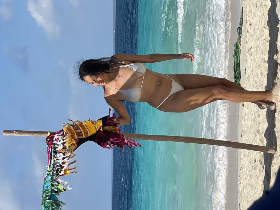 The scene depicts a sunny beach environment in Zanzibar, Tanzania. A lone female individual stands in a three-quarter profile facing the viewer's right, smiling. She is positioned slightly to the right of a vertical wooden pole rooted in the white sand, with her left hand resting on the pole. She wears a light grey two-piece bikini, a navel piercing, and a necklace with a circular pendant. Her dark hair appears wet and is pushed back from her face.
Several colorful, patterned fabrics, including items with fringe and tropical motifs, are draped over the upper portion of the wooden pole, extending horizontally to the left. The background features turquoise and deep blue ocean waters with white waves breaking on the shore. The sky is clear blue with sparse white clouds. Distant boats or landmasses are faintly visible on the horizon line over the ocean. Shadows are cast on the sand, indicating bright sunlight. A dark green rope or netting is partially visible on the sand in the lower right quadrant, and a portion of a blue object is in the bottom left foreground.