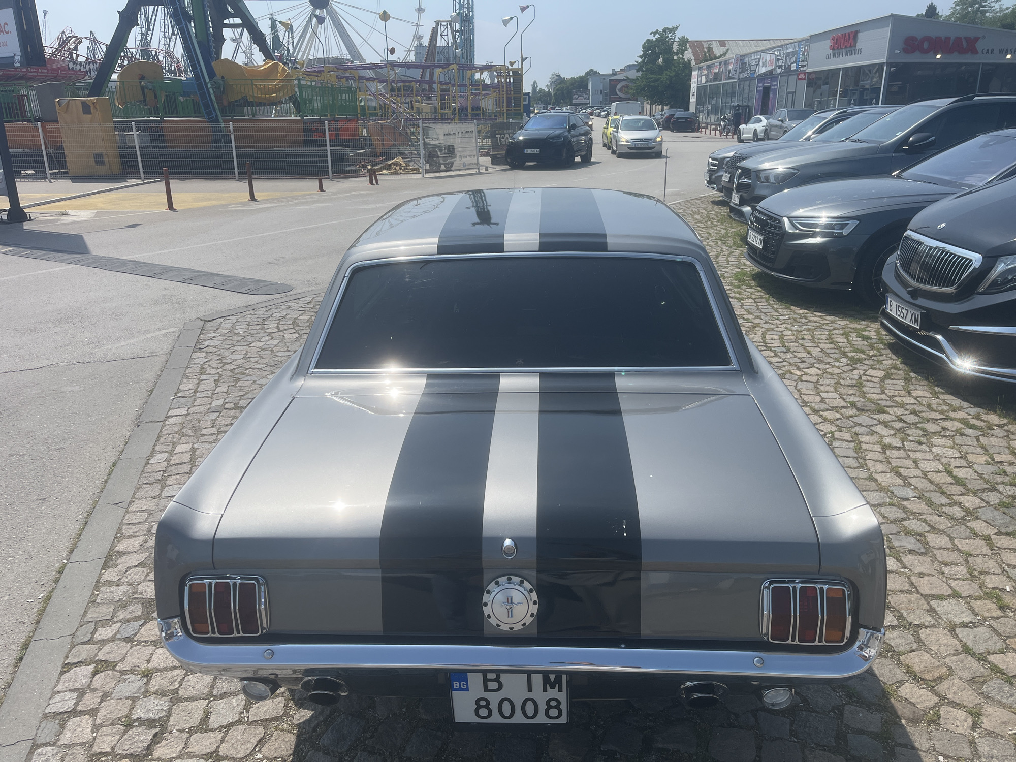 The image captures the rear view of a classic grey Ford Mustang with prominent black and lighter grey racing stripes, parked on a cobblestone surface. The car features a chrome bumper, triple vertical taillights, dual exhaust pipes, and a circular fuel cap adorned with a horse emblem. Its Bulgarian license plate reads "BG B IM 8008".
To the left of the car, a paved road with multiple speed bumps leads towards an amusement park, identifiable by large Ferris wheel structures and other ride components behind a mesh fence. On the right, a row of modern, dark-colored cars, including SUVs and sedans, are parked on a similar cobblestone area in front of a commercial building. The building features signage for "SONAX CAR WASH & DETAILING".
The scene is set in an urban environment, under bright, clear daylight, casting distinct shadows and making the car's paint glisten. The clear blue sky is visible above. The numerous vehicles and commercial establishments suggest a bustling area. Other visible Bulgarian license plates include "B 1542 XA", "B 2212 XM", and "B 1557 XM". Partial text "AC." and "002" are also visible on a sign near the amusement park. The location is Varna, Bulgaria.