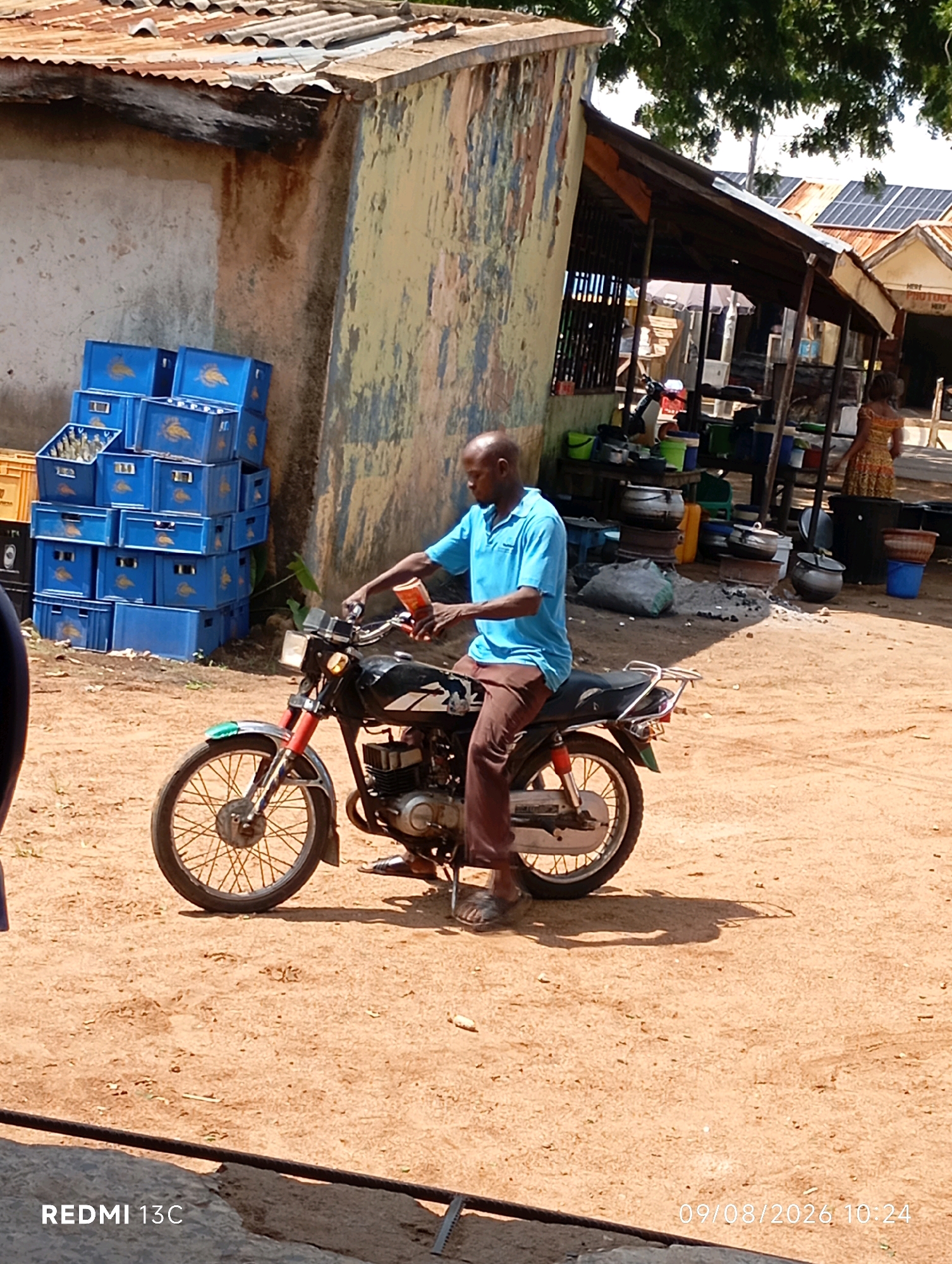 In the city of Shendam, Nigeria, a bald man in a light blue t-shirt and brown pants is seated on a black motorcycle. He holds a red bottle in his right hand. The motorcycle is parked on a dirt surface. In the background, stacks of blue plastic crates, some filled with bottles, are visible against a weathered, textured wall. A covered outdoor market area with a sloped tin roof, supported by wooden posts, is situated further back. Several cooking pots and buckets are present in this market area. Solar panels are affixed to another roof in the distance. The scene is illuminated by daylight, casting shadows on the ground.