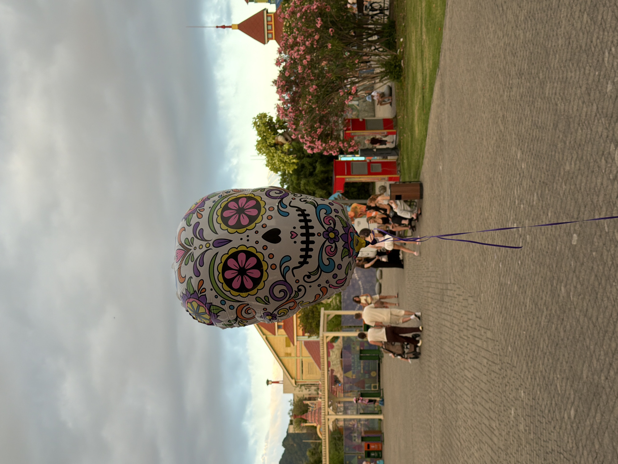 A large, round balloon decorated with a colorful sugar skull design floats in the sky against a cloudy, overcast sky. The balloon is tethered by a thin purple ribbon that stretches across the frame. In the background, a paved walkway curves through a park-like setting. Beyond the walkway, there are buildings with red roofs, a vibrant pink flowering bush, and green trees. Several people are visible in the background, some sitting on benches, others walking. One person appears to be pushing a stroller. The overall scene suggests a casual outdoor gathering or a visit to a park or amusement area. The lighting indicates it might be late afternoon or early evening. There is no visible text in the image.