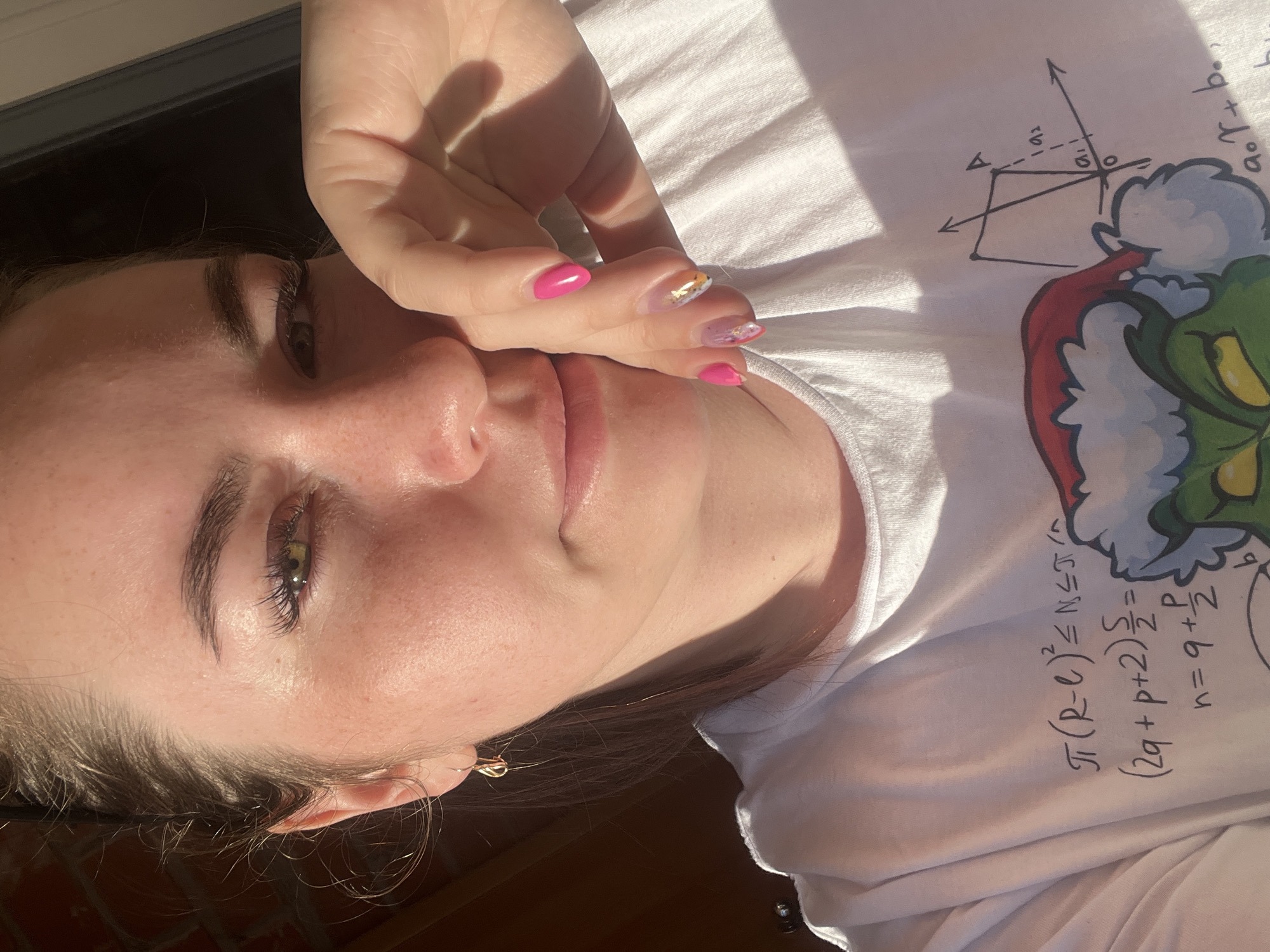 A young woman is captured in a close-up, eye-level shot, with strong, uneven lighting suggesting direct sunlight. She is positioned with her head tilted, looking towards the viewer, and her right hand is brought up to partially cover the lower left side of her face, extending from her nose to her chin. Her fingernails are painted with a vibrant pink polish, and some nails feature additional gold and iridescent glitter designs. A gold hoop earring is visible in her left ear.
She is wearing a white T-shirt that features several graphical elements. On the shirt, there's a cartoon illustration of the Grinch wearing a red Santa hat, positioned towards the right side of the frame. To the left of the Grinch, mathematical equations and a diagram are visible. The diagram includes a coordinate system with labeled vectors "a1" and "a2", forming a geometric shape. Text snippets on the shirt include "a.r + b." and equations such as "П(R-C)²=NSTA", "(2q+p+2)/2 =", "P", and "n = 9 + ½".
The woman has dark eyebrows and long eyelashes, complementing her brown eyes. Her skin shows some freckles and is highlighted by the bright sunlight, which also casts prominent shadows on her hand and shirt. Her dark hair is pulled back, with some strands framing her face. The background is dark and out of focus. The image was captured in Chaikovsky, Russia, during what appears to be a sunny day, with bright sun creating a high-contrast scene.