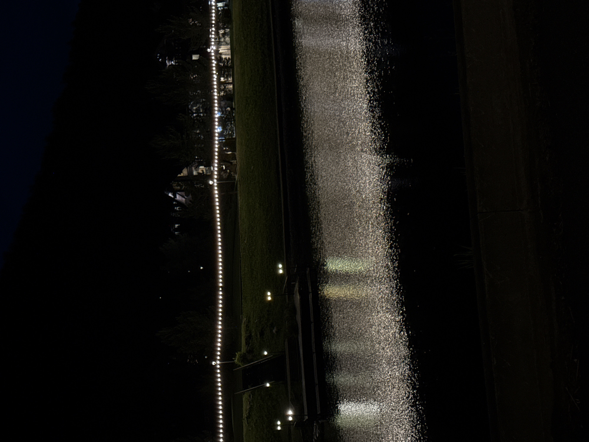 The image depicts a nighttime scene along a body of water, likely a river or lake. In the foreground, the dark water reflects scattered lights, creating a shimmering effect. Along the bank, a set of concrete steps leads up to a grassy area. Several lampposts with multiple bulbs are visible along the bank, casting yellow and white light. A string of small, white lights runs horizontally along the top of the grassy area, above a line of silhouetted trees. In the background, the dark shapes of buildings and a sloping hillside are faintly visible against the night sky. The overall impression is one of a peaceful, possibly resort-like, setting at night. There are no people visible in the image. The scene is not identifiable as being specifically in Urlu-Aspak, Russia, based solely on the visual information provided.