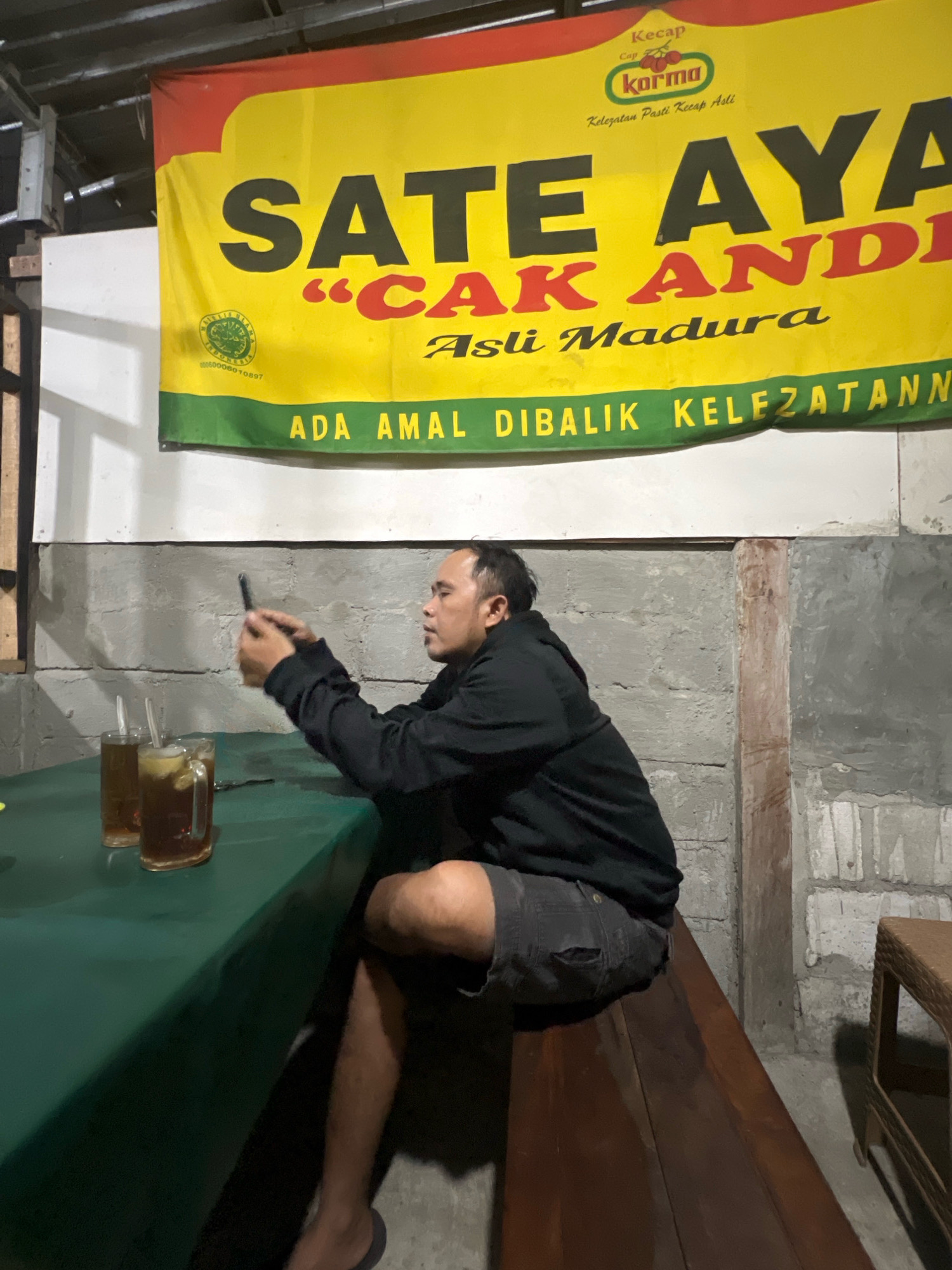 A man is sitting on a wooden bench in front of a green table, holding a phone in his right hand. On the table are two glasses of iced tea. Behind the table is a yellow banner with red and green lettering. The banner reads "Sate Aya" in large black letters, with "Cak Andi" in red letters below it. There is also text in Indonesian that translates to "Asli Madura" (Original Madura) and "Ada Amal Dibalik Kelezatann" (There is Charity Behind the Deliciousness). The background consists of a grey, rough-textured wall. The scene appears to be outdoors, possibly at a food stall or restaurant in Trihanggo, Indonesia, given the presence of the banner advertising "Sate Ayam Cak Andi Asli Madura" and the general setting.