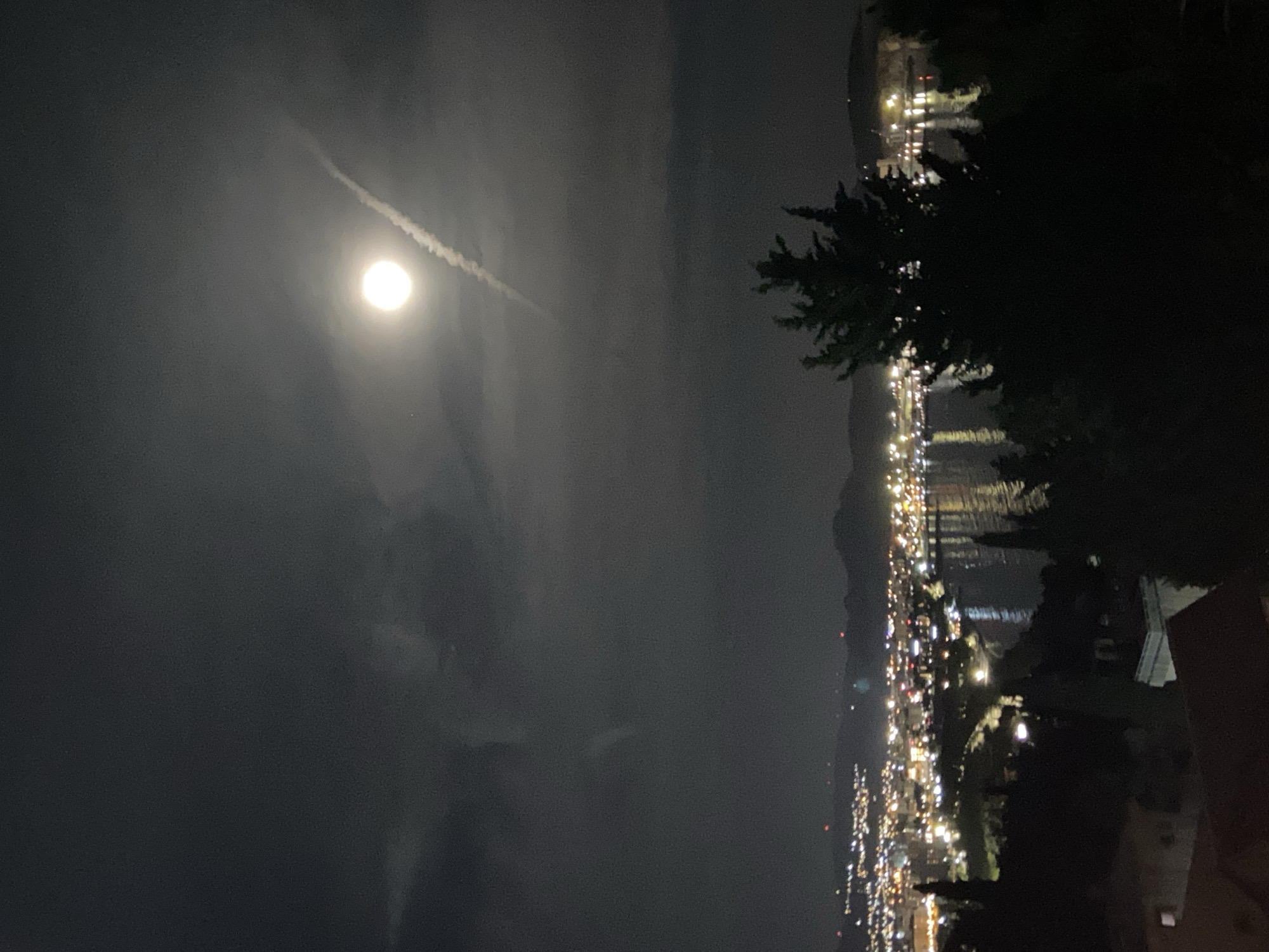 The image depicts a nighttime scene in Šušanj, Montenegro. A bright moon is visible in the sky, partially obscured by clouds. A faint contrail stretches out from the moon. The lights of the city line the coast, reflected in the water. Dark trees and vegetation are silhouetted in the foreground.