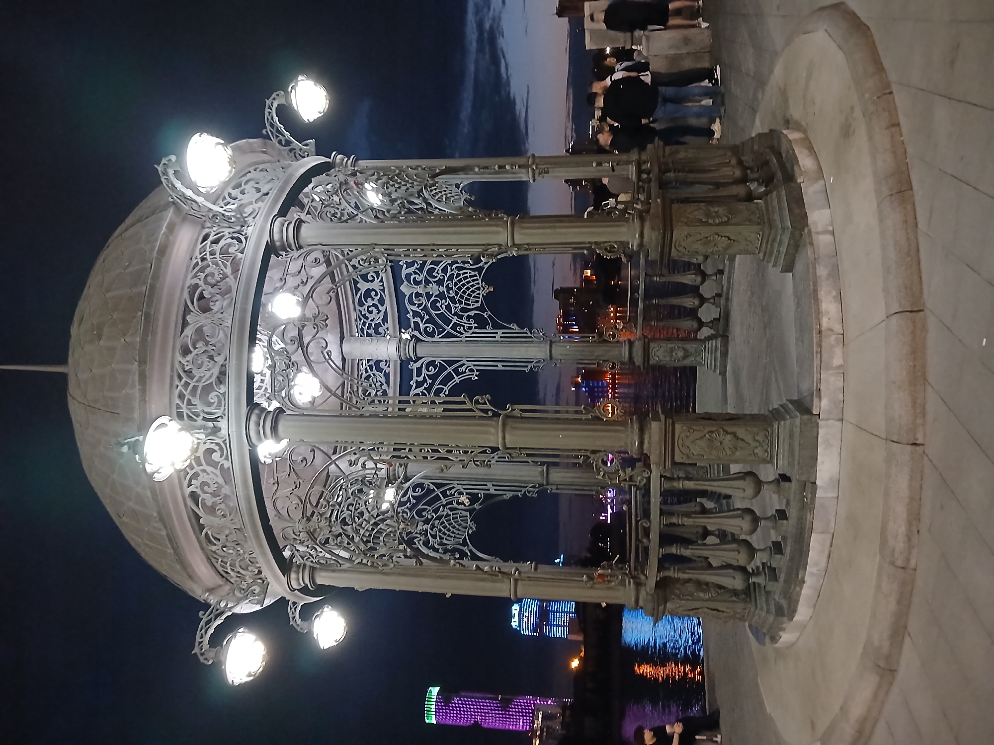 The image shows the Observation Deck "The Ring" located on the embankment of the Iset River in Yekaterinburg, Russia, at dusk or night. The main subject is a decorative gazebo made of intricate metalwork, illuminated by several lamps. In the background, the Iset River reflects the lights of the city, including illuminated skyscrapers and buildings. Several people are visible near the gazebo and along the embankment. One person is sitting on the edge of the embankment to the left, and a group of people is standing near the gazebo on the right. The sky is dark with some clouds visible. The ground is paved with light-colored tiles. The overall scene depicts a vibrant urban waterfront area at night.