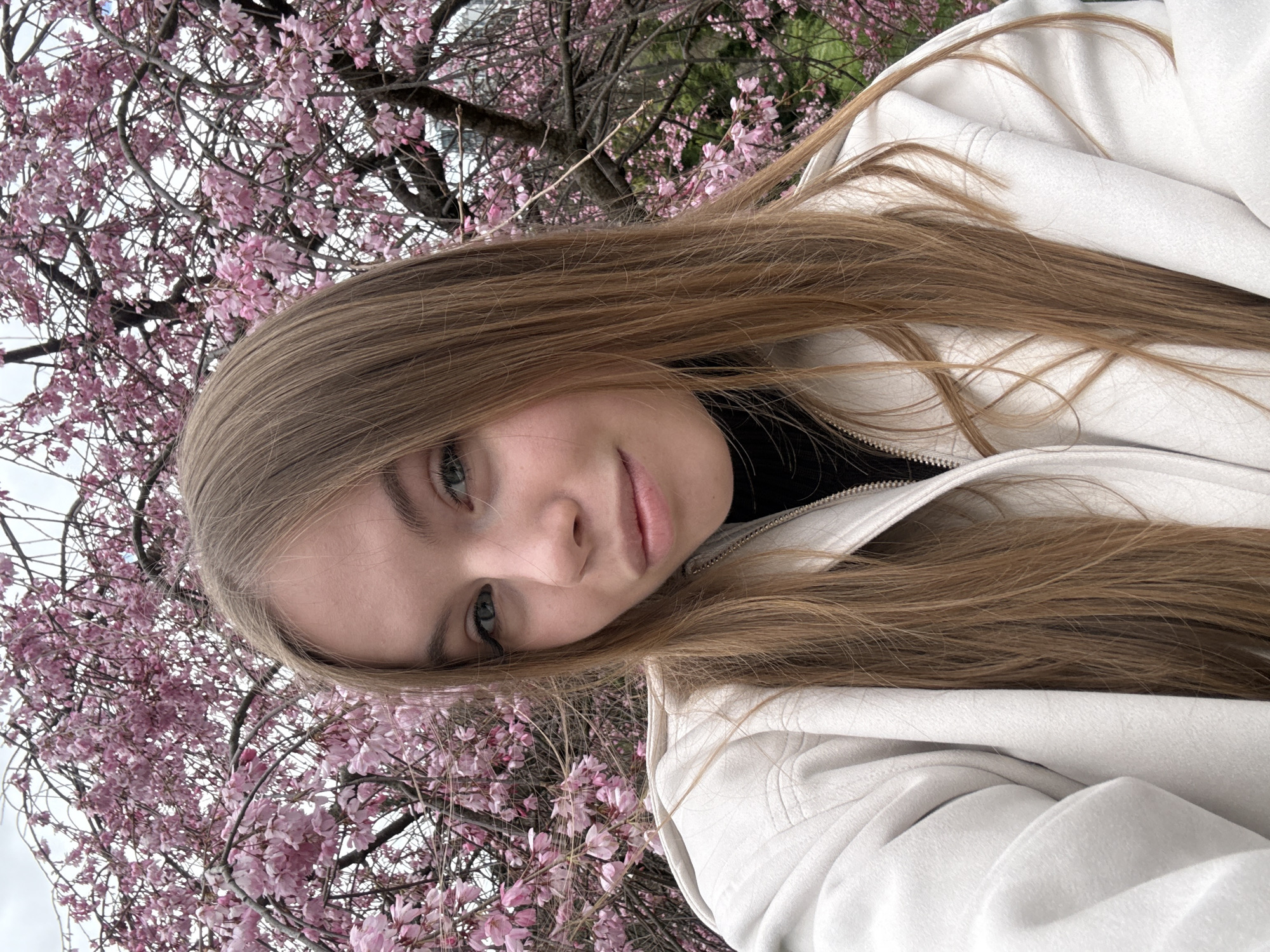 A young female subject is depicted in a close-up selfie-style portrait, positioned against a background dominated by a blooming weeping cherry blossom tree in Sochi, Russia. The subject has long, straight, light brown hair, light-colored eyes, and fair skin, looking directly at the camera with a slight, neutral expression. She is wearing an off-white or light beige jacket or coat over a black turtleneck garment, visible at the neckline. The jacket features a zipper closure. The background consists primarily of a dense canopy of pink cherry blossoms and dark tree branches against an overcast sky. Green foliage is minimally visible at the lower right.