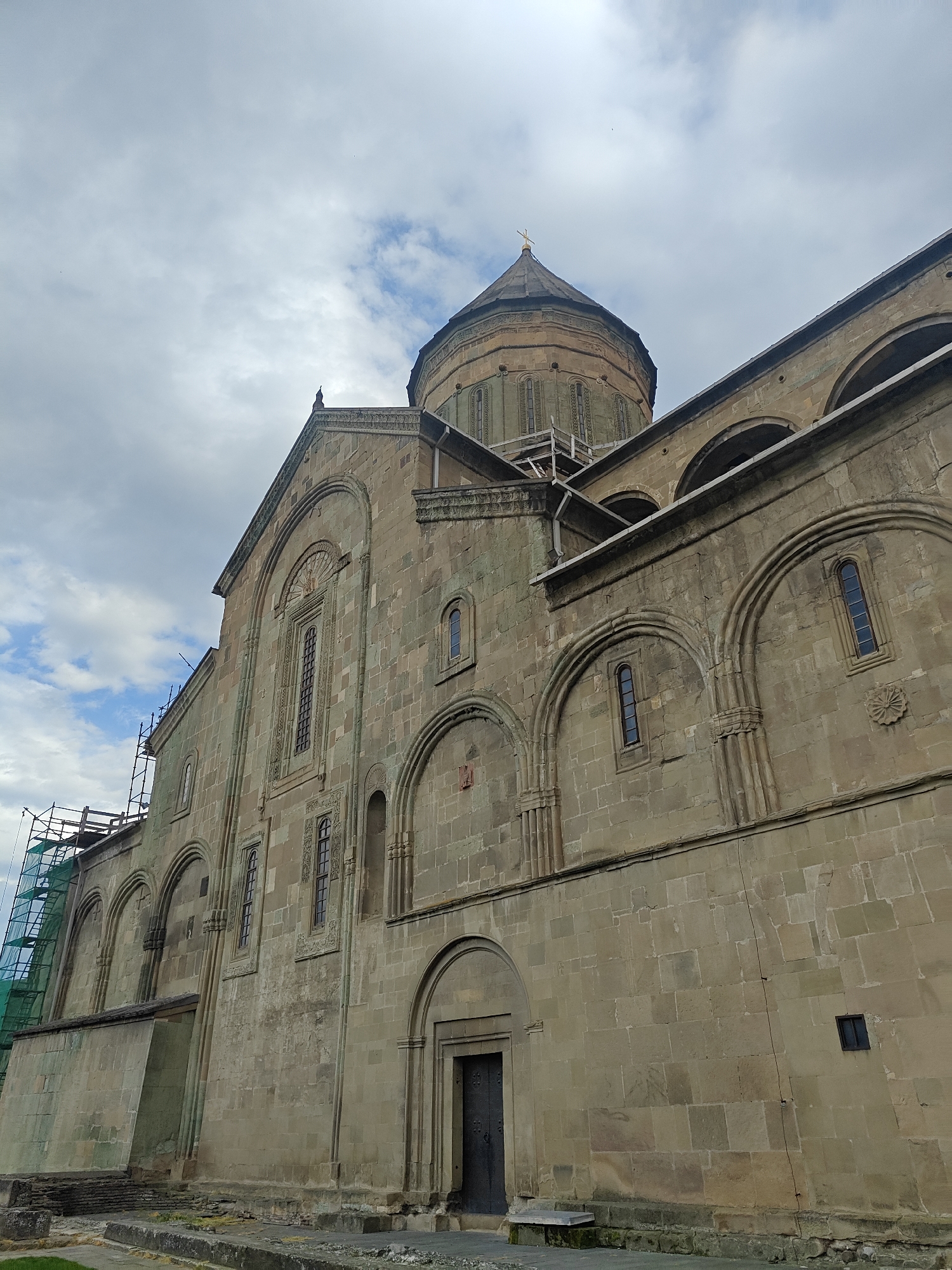 A low-angle shot captures a large, ancient stone church or cathedral under a cloudy sky. The building is constructed from weathered, light brown and greenish-gray stone blocks, giving it a venerable appearance. A prominent, multi-faceted dome, topped with a small golden cross, dominates the upper section of the structure, reaching into the sky.
The facade features numerous architectural details, including tall, narrow arched windows with ornate stone carvings, and several larger decorative arches along the upper walls. One dark, arched wooden door is visible at the base of the building on the right side. On the far left, a substantial section of scaffolding, covered with a bright green mesh netting, is erected against the building, indicating ongoing renovation or maintenance work. The sky is mostly overcast with diffuse white and gray clouds, though some patches of blue sky are visible. No people or visible text are present in the image.