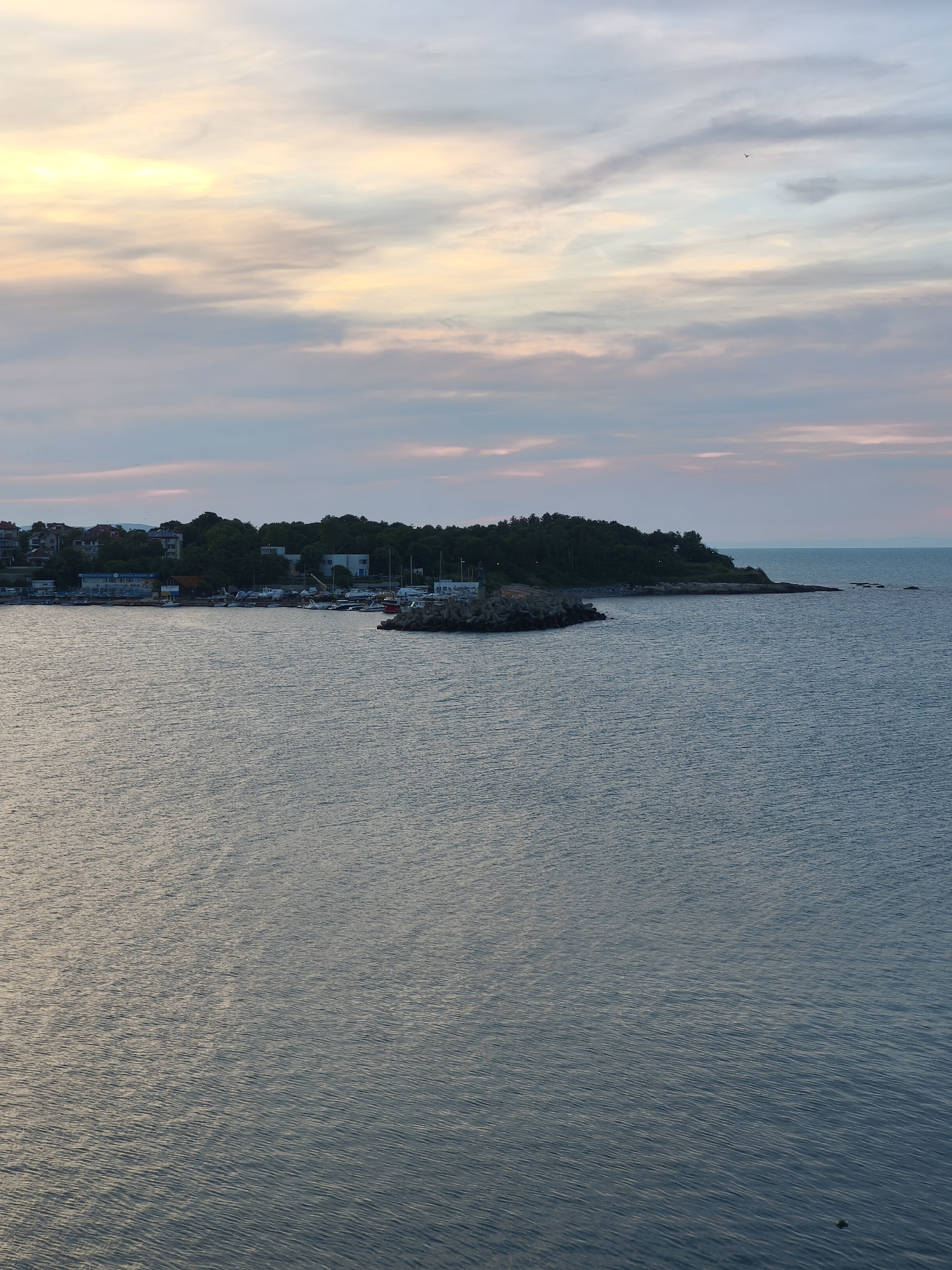 The image shows a coastal view of Tsarevo, Bulgaria, likely during a sunset. The foreground is dominated by the calm, blue-gray waters of the sea, reflecting the soft light of the sky. In the middle ground, a rocky coastline juts out into the water, lined with a dense green forest. Along the shoreline, a small harbor is visible, with several boats moored. Beyond the harbor, a cluster of buildings, likely residences and businesses, are nestled amongst the trees. The background features a sky filled with wispy clouds painted in hues of pale yellow, pink, and blue, indicating the setting of the sun. The overall atmosphere is serene and peaceful.