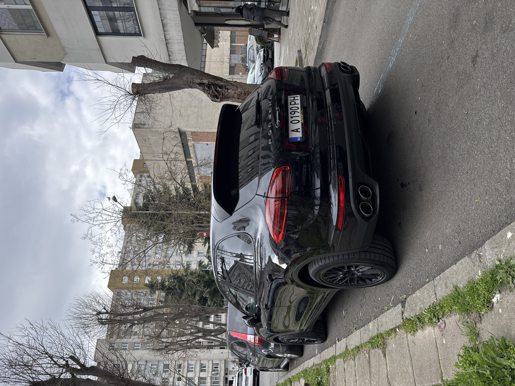 The image shows a black Porsche Cayenne S parked on a street in Burgas, Bulgaria. The car has a Bulgarian license plate A 0190 PH. The location is urban with buildings and trees in the background. A person is standing in front of a cafe. The car is parked on the side of a road near a sidewalk.
