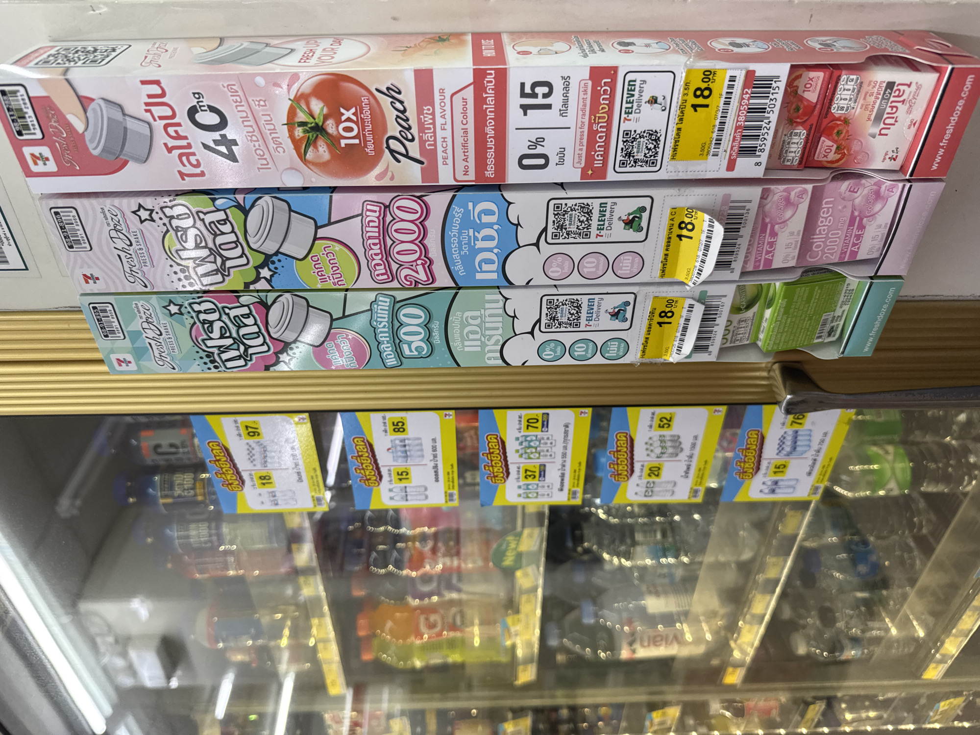 The image captures a close-up view of a refrigerated display case filled with various drinks and products, likely in a convenience store. The display case is located at a 7-Eleven store in Pattaya City, Thailand, as indicated by the prominent 7-Eleven branding and Thai text on the product packaging and promotional signs.
The focus is on the products mounted on the side of the display, which are primarily health and beauty drinks. These products are displayed in colorful packaging with Thai text and imagery, including illustrations of fruits and caps. Several promotional tags, often featuring discounts and product information in Thai, are attached to the products and the display itself.
Inside the refrigerated section, numerous bottles of water and other beverages are visible, some with labels showing Thai text and prices. The lighting within the display case creates reflections on the glass. The overall scene is that of a retail environment, highlighting the product offerings of a 7-Eleven store in Pattaya.