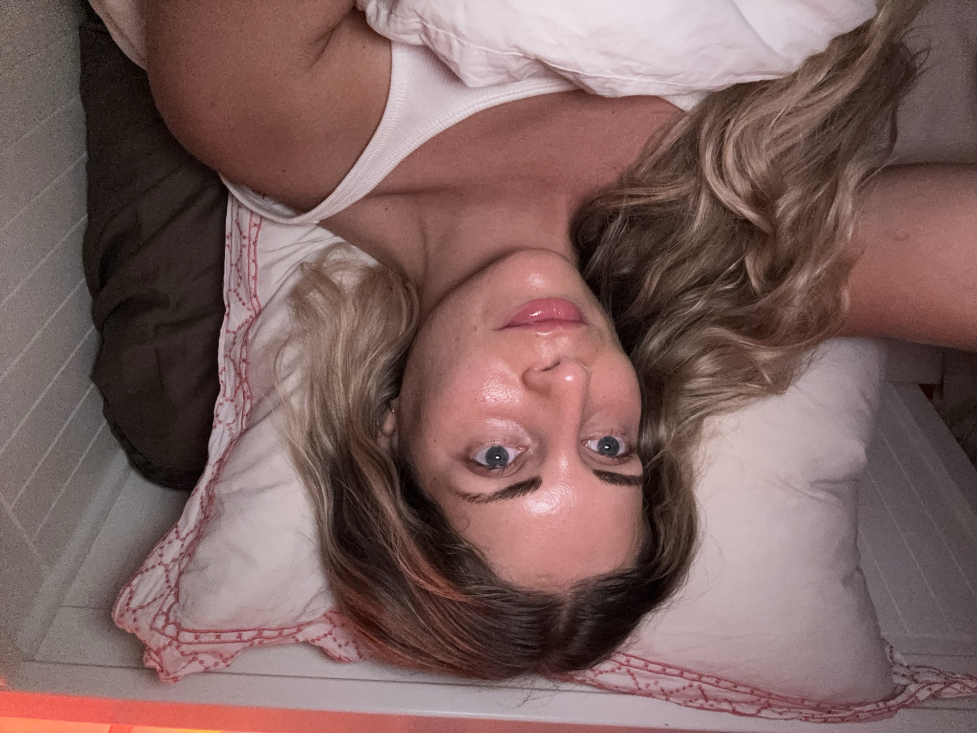 A woman with blonde hair, wearing a white tank top, lies in bed, partially covered by a white duvet. She is looking at the camera. The bed has a white pillow with red trim. The background appears to be a wall with vertical wooden planks. The lighting in the room is warm and appears to be coming from above, casting a reddish hue on the top of the image. The scene is a bedroom.