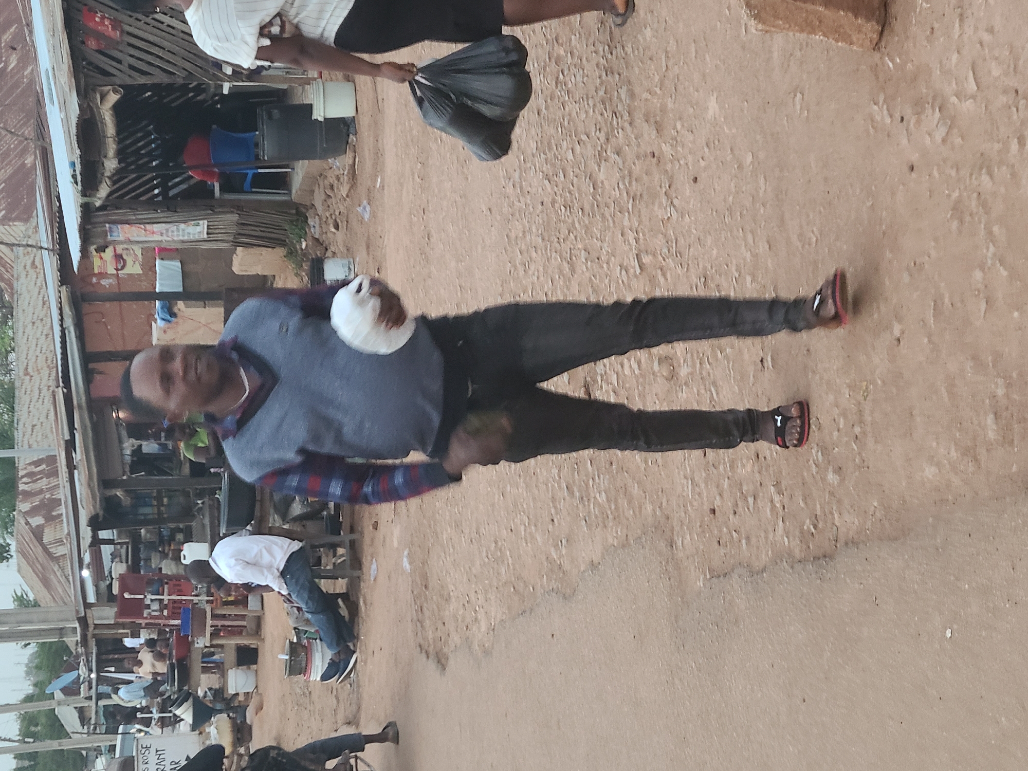 The image depicts an outdoor scene in Jos, Nigeria, characterized by a dusty, unpaved road and informal commercial structures on the left side of the frame. A man is centrally positioned, wearing a blue and red checkered shirt, a grey vest, black pants, and sandals with red accents. He holds a white container or object in his left hand.
In the background, a collection of stalls or small shops constructed from corrugated metal and wood are visible. These structures appear to house various vendors, with plastic chairs and other items indicating commercial activity. Several people are present near the stalls, engaged in what appears to be casual interactions or business transactions.
Another man is slightly visible, carrying a black bag. The road surface is uneven and worn, indicative of heavy pedestrian and vehicular traffic. The overall environment suggests a bustling, informal commercial district in Jos, Nigeria.