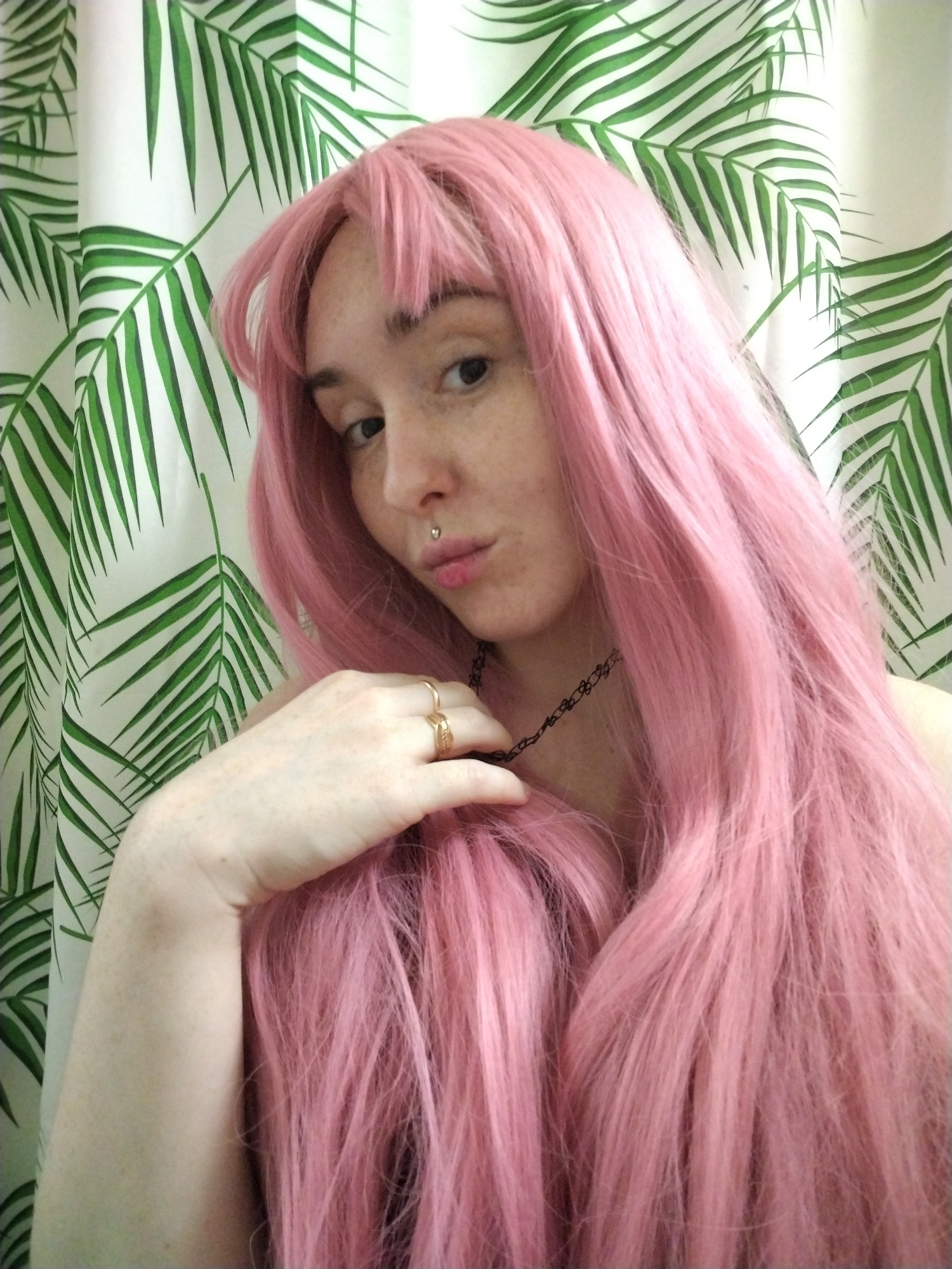 A fair-skinned person with freckles is posing for a selfie. They have pink hair with bangs and are wearing a black choker necklace and rings. The background is a shower curtain with a green palm leaf print. There is no visible information to determine the location is in Izhevsk, Russia.