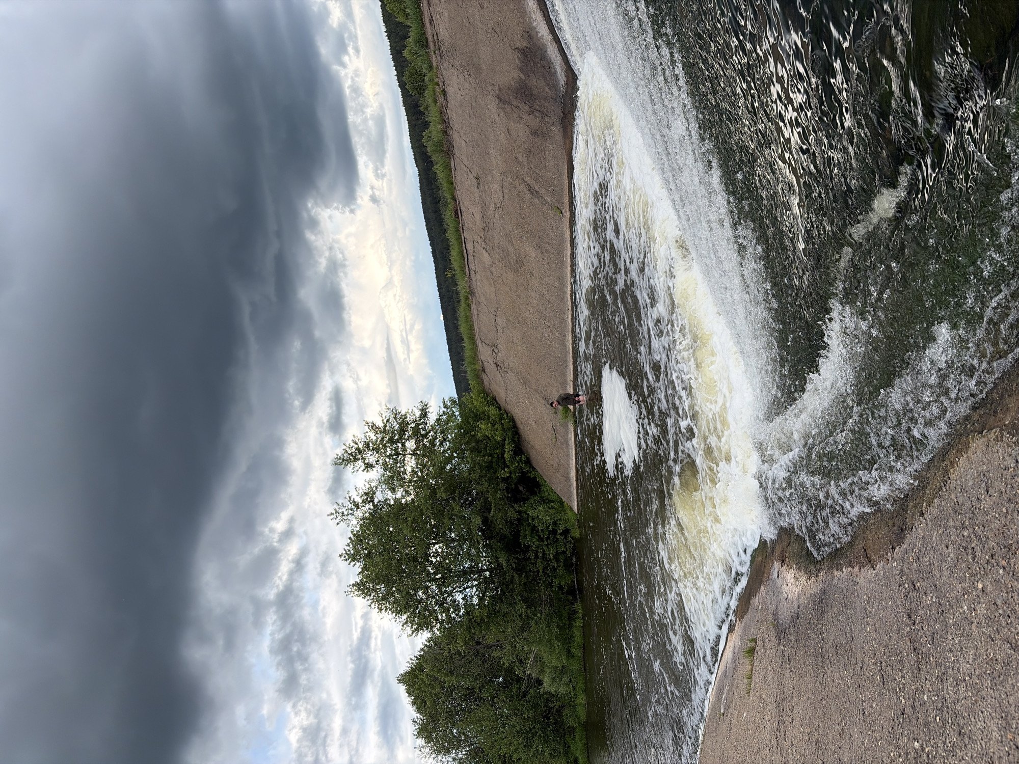 The image shows a waterfall or dam in Nizhnii Vishur, Russia. Water cascades down a concrete structure into a pool below. There's a person standing in the pool near the base of the waterfall. Lush green trees are visible along the left side of the concrete structure. The sky is dominated by ominous dark storm clouds.