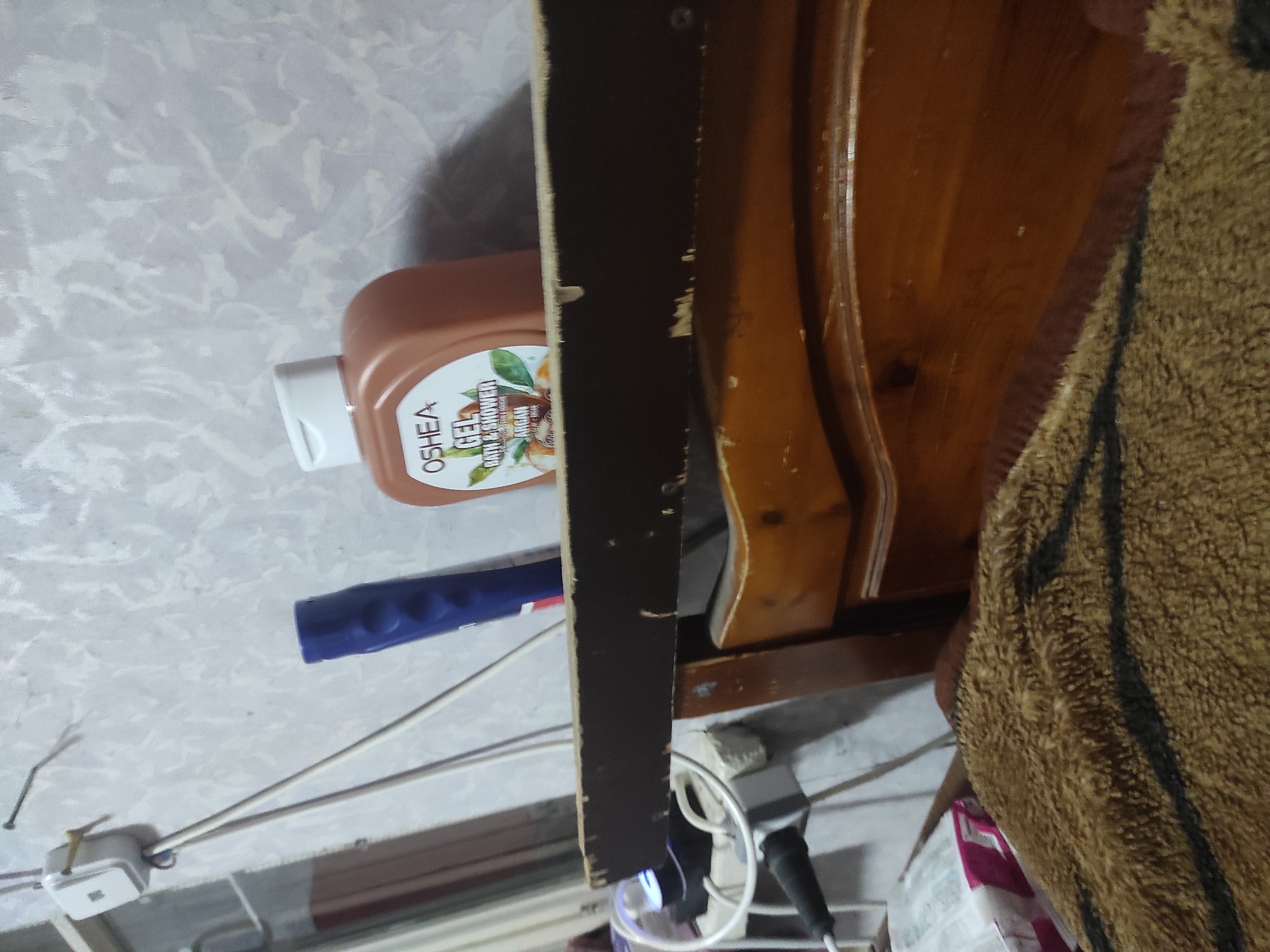 The image shows a close-up, slightly upward view of a bedside shelf in what appears to be a bedroom. On the shelf, there are two toiletries: a brown bottle of "OSHEA GEL BATH & SHOWER" with an Argan oil label, and a blue tube, likely another toiletry item. To the left of the shelf, a white electrical outlet is mounted on a textured, light-colored wall. White and grey wires are connected to the outlet and run down. Below the shelf, the top of a wooden headboard is visible, with a brown, fuzzy blanket draped over it. The overall scene suggests a functional, lived-in space, possibly a bedroom or a dorm room.