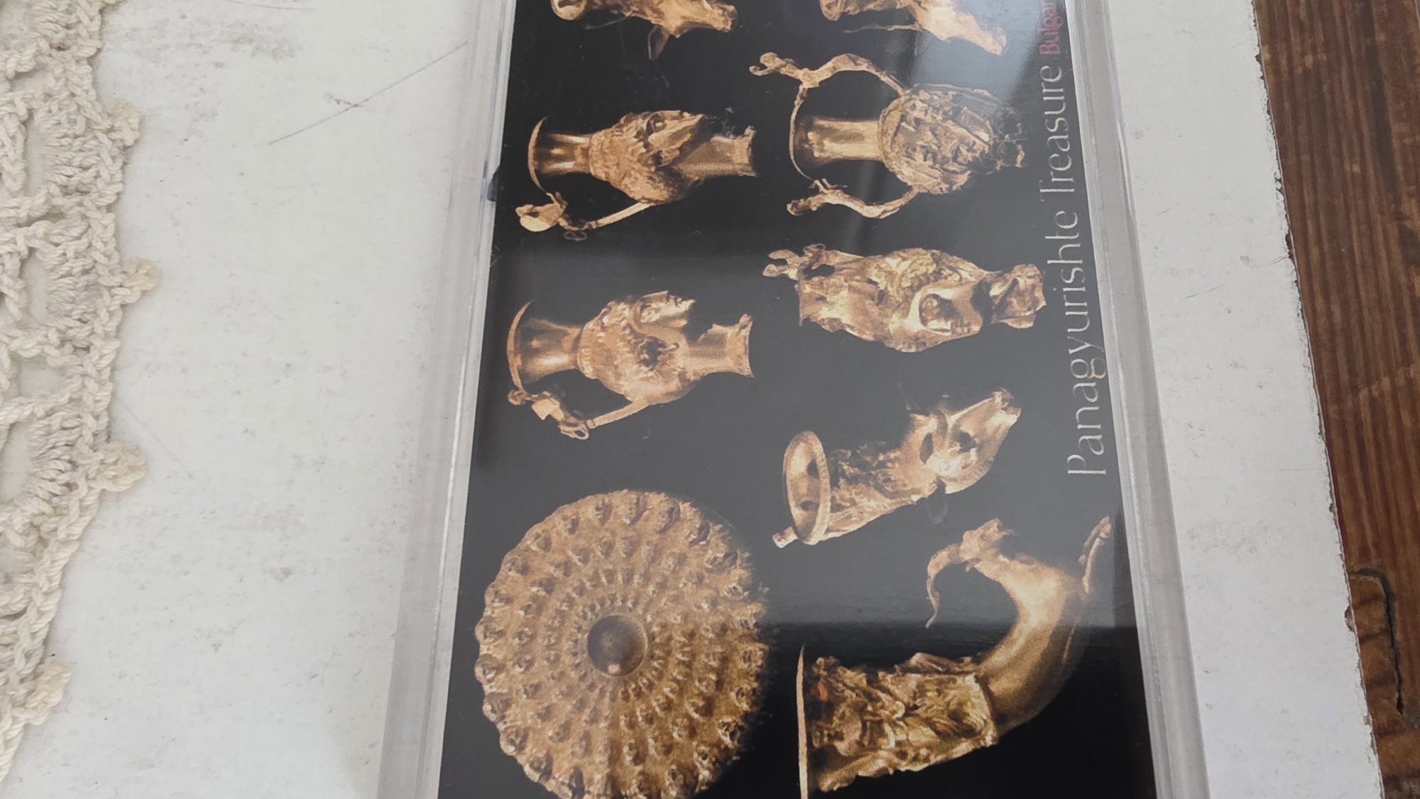 The image shows a postcard or a photograph of the Panagyurishte Treasure, found in Bulgaria. The card features a black background showcasing various golden artifacts, including rhytons shaped like animal heads and other decorative pieces. The text "Panagyurishte Treasure Bulgaria" is visible on the card. The card is encased in a clear plastic sleeve and placed on a surface that includes a beige crochet lace object on one side and a dark wooden surface on the other. The location of the Panagyurishte Treasure find is near the town of Panagyurishte, not in Blagoevgrad.