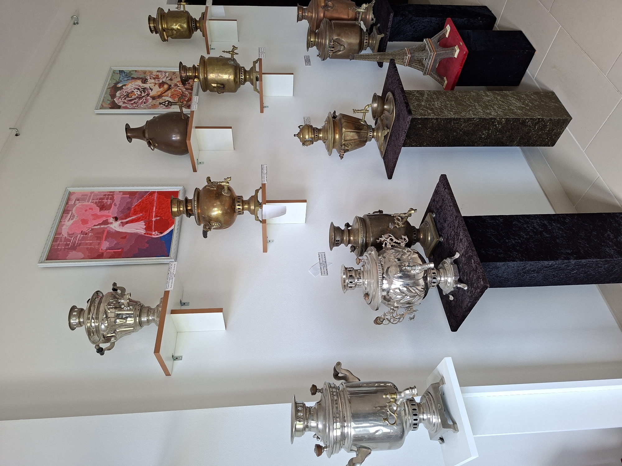 This image displays a collection of antique samovars and decorative vases arranged on shelves and display stands against a white wall. The room appears to be a museum or exhibition space. On the wall, there are two framed artworks; one features a stylized depiction of a dancer in red and pink, and the other is a portrait of a woman adorned with flowers. The samovars vary in size, material, and design, with some made of polished metal and others of brass. There are also several decorative vases, including a large dark brown one. In the foreground, on the right side, a miniature replica of the Eiffel Tower sits on a pedestal. The floor is tiled with light-colored squares. The lighting suggests an indoor environment, likely with artificial illumination. The scene depicts a static display of historical artifacts. Based on available information, this collection is housed at the Ialoveni Museum of History and Ethnography in Ialoveni, Moldova.