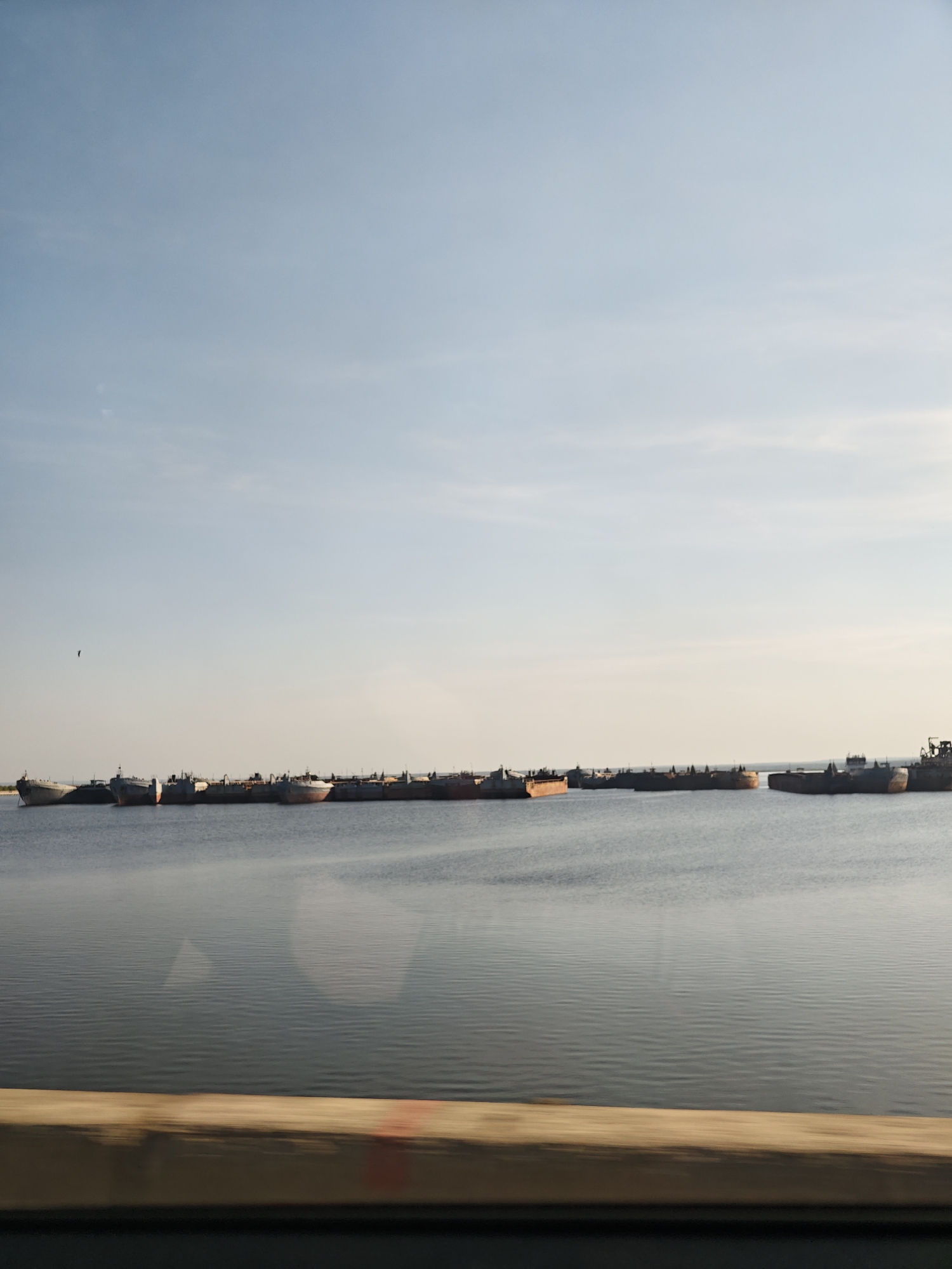 The image depicts several barges on the water in Chaikovsky, Russia. The sky is light blue with some clouds. A bird is flying in the sky. A barrier or rail is visible in the foreground.