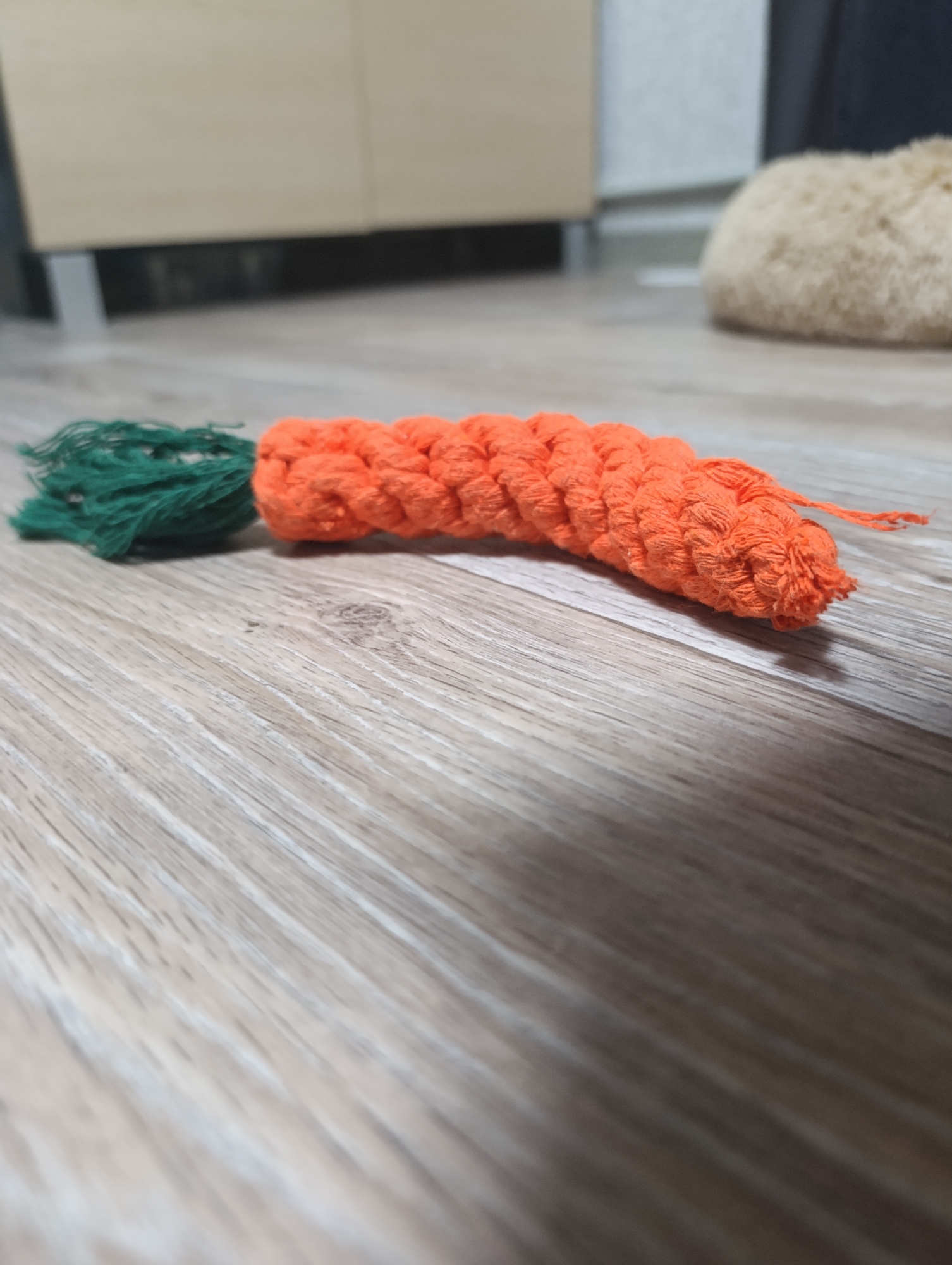 An orange crocheted carrot with a green yarn stem lies on a light brown wooden floor. The carrot is oriented horizontally, with the green stem to the left. The background shows a blurry, light-colored cabinet and a fuzzy, beige object. The scene is indoors in an environment that appears to be a home.
