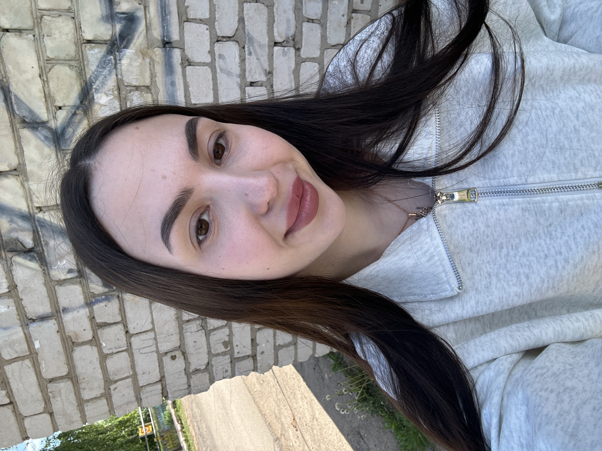 A young woman with dark hair poses for a selfie in Фоки, Russia. The woman is wearing a light gray hooded sweatshirt with a zipper. Behind her is a white brick wall with some graffiti on it. A concrete walkway is visible with greenery to the side, suggesting an outdoor location within the city.