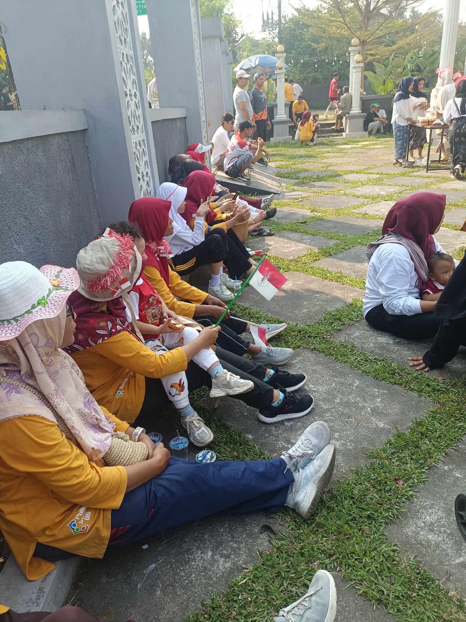 In the city of Sleman, Indonesia, a group of people are seated on the ground in front of a gray concrete wall with ornate white decorative panels. The ground is a mixture of paved stones and patchy grass. Many of the individuals are wearing traditional Indonesian attire, including headscarves (hijabs) in various colors like red, maroon, and white. Some are dressed in yellow shirts, and one person is wearing a white shirt. Several children are present, some holding small Indonesian flags (red and white). One child is being held by an adult. In the background, there are more people standing and walking, some gathered around tables, suggesting an event or gathering. The overall scene appears to be an outdoor public space, possibly a park or a plaza, during daylight hours.