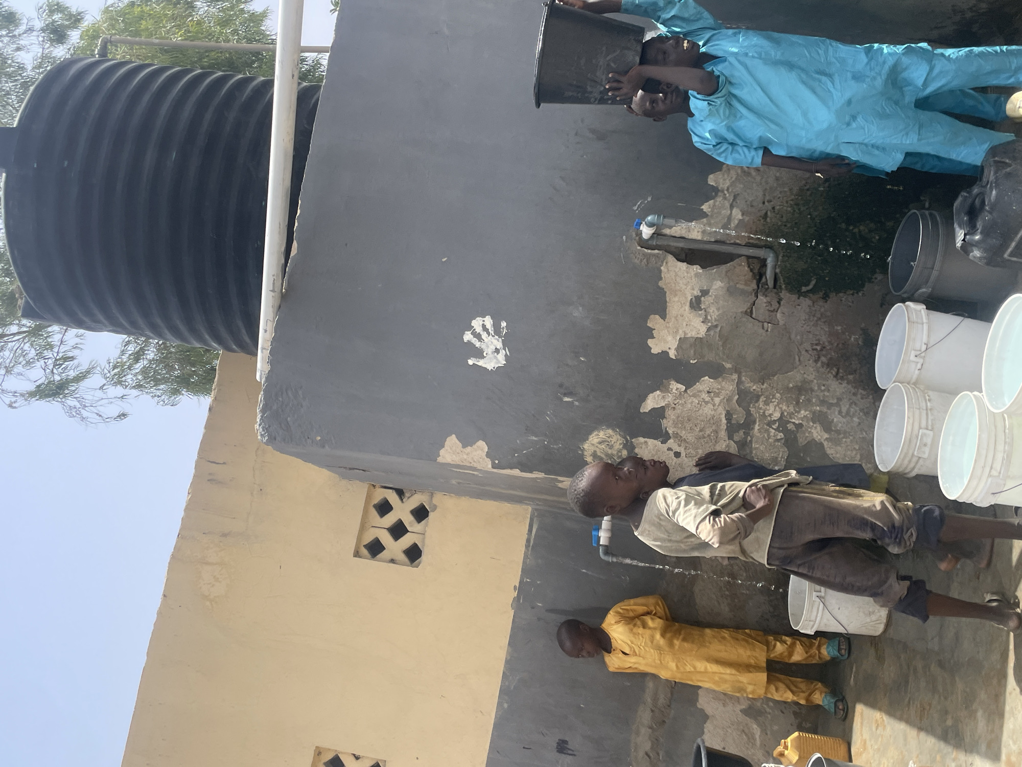 In an outdoor setting in Unknown, Unknown, several individuals are interacting with a water source near a building.
A large, black, corrugated water tank stands adjacent to a light yellow building and a taller, gray structure. The gray structure has an exterior water faucet with water flowing from it. A handprint is visible on the gray wall.
Four young people appear to be collecting water. One wears a light blue tunic and fills a dark container. A second person, wearing a light tan top and darker pants, stands near the flowing water. A third wears a yellow tunic. Several white buckets are on the ground.
The scene is illuminated by sunlight with a clear blue sky visible.
