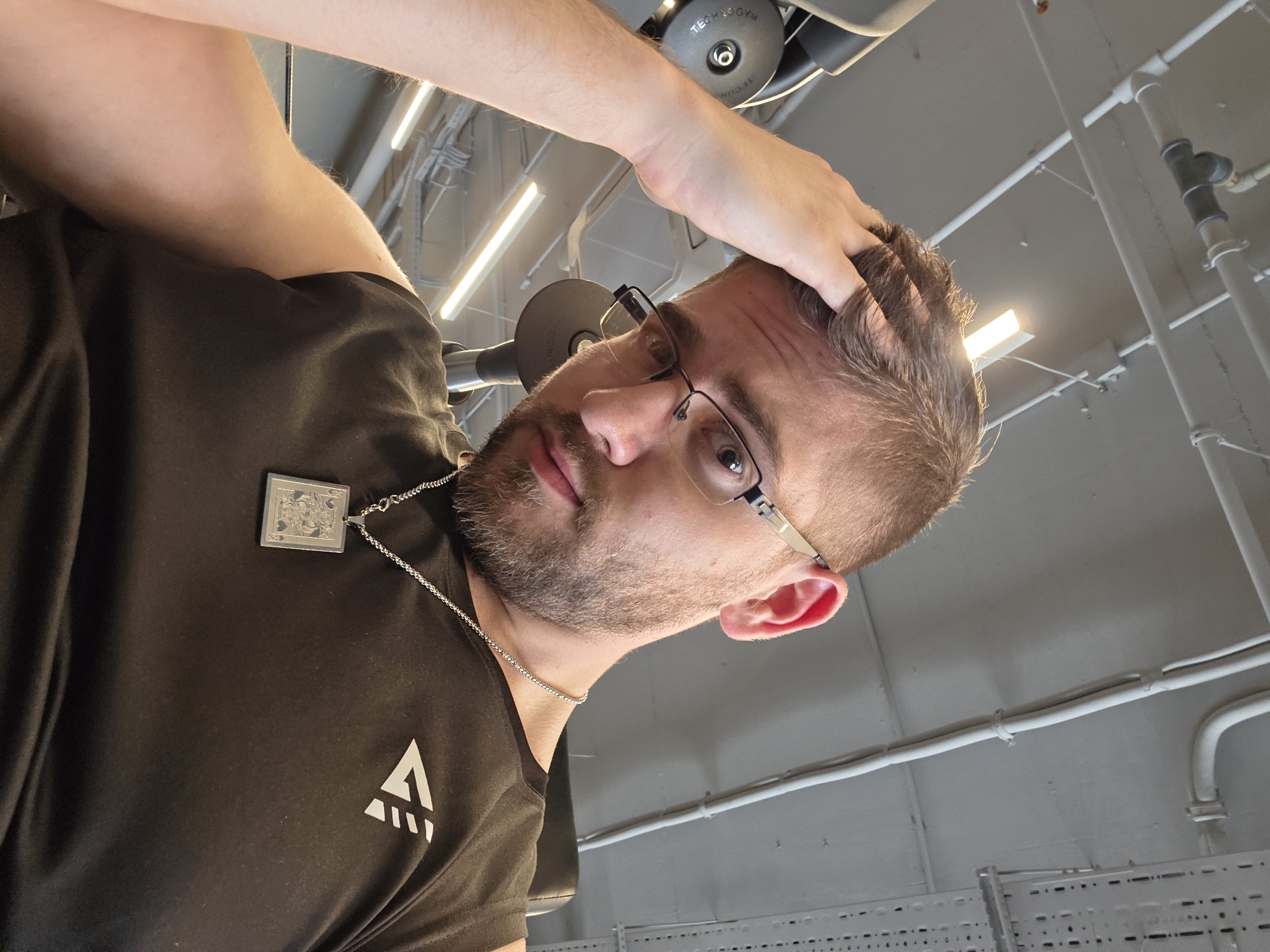 This media file presents a portrait of a man in what appears to be an indoor gym in Plovdiv, Bulgaria. The man, likely in his 20s or 30s, has short brown hair and a light beard. He wears black-framed glasses and a dark, short-sleeved t-shirt with a white, stylized triangular logo resembling an upward-pointing arrow or the letter "A" on the left chest. Around his neck, he sports a silver chain from which hangs a rectangular pendant, clearly depicting the Queen of Spades playing card. His right hand is casually placed in his hair, and he looks directly at the camera with a neutral expression.
The background provides clear cues to the setting. Visible gym equipment includes a dark gray pulley wheel with the brand name "TECHNOGYM" inscribed on it. The ceiling area reveals industrial-style fluorescent strip lighting, white pipes, and conduits, indicative of a functional and well-lit commercial space like a fitness center. The overall lighting is bright and even, suggesting either a well-lit daytime interior or a facility with robust artificial illumination. The activity captured is a posed photograph within this gym environment.